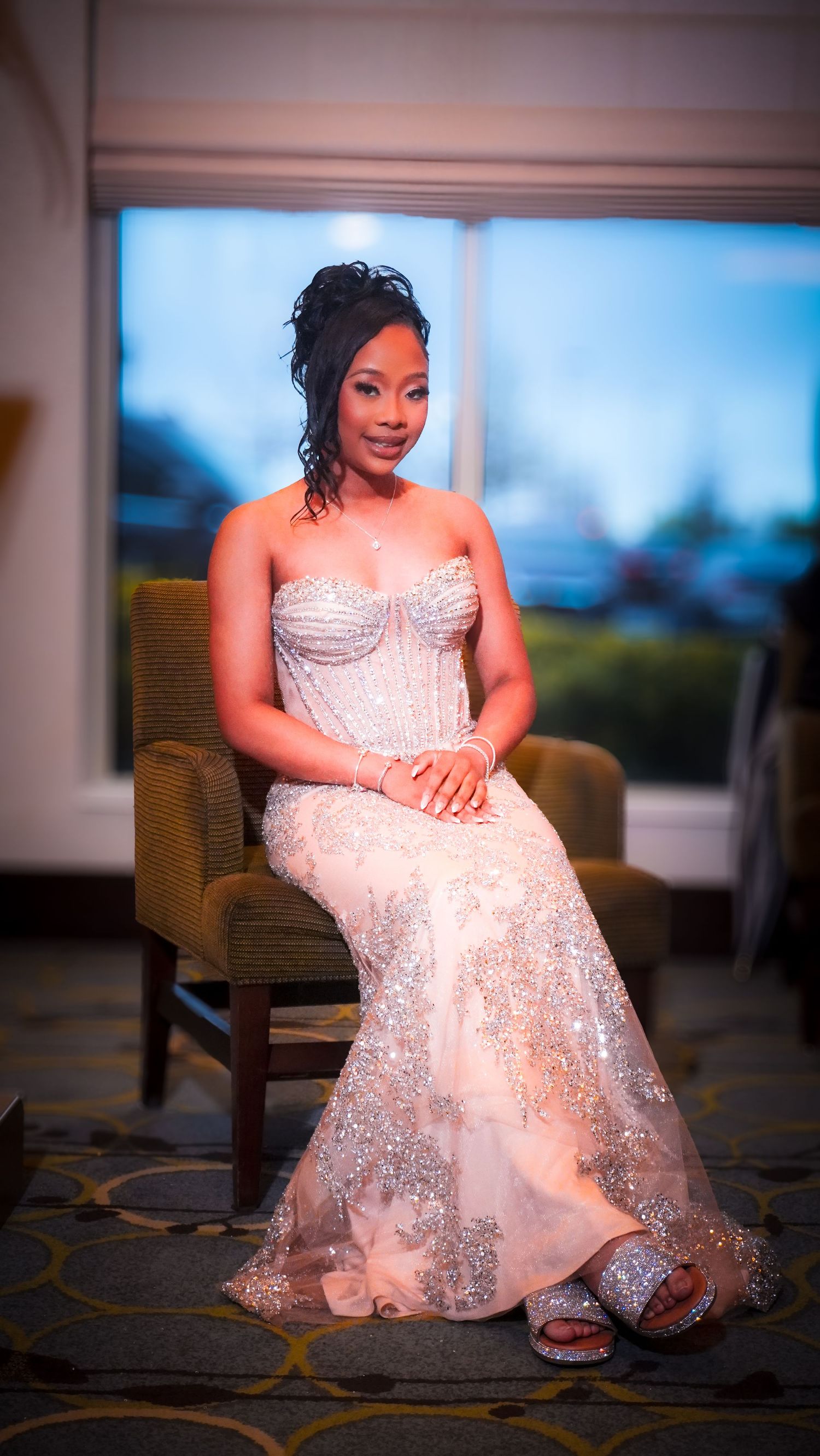Woman in an elegant silver evening gown seated indoors, formal portrait with soft lighting.