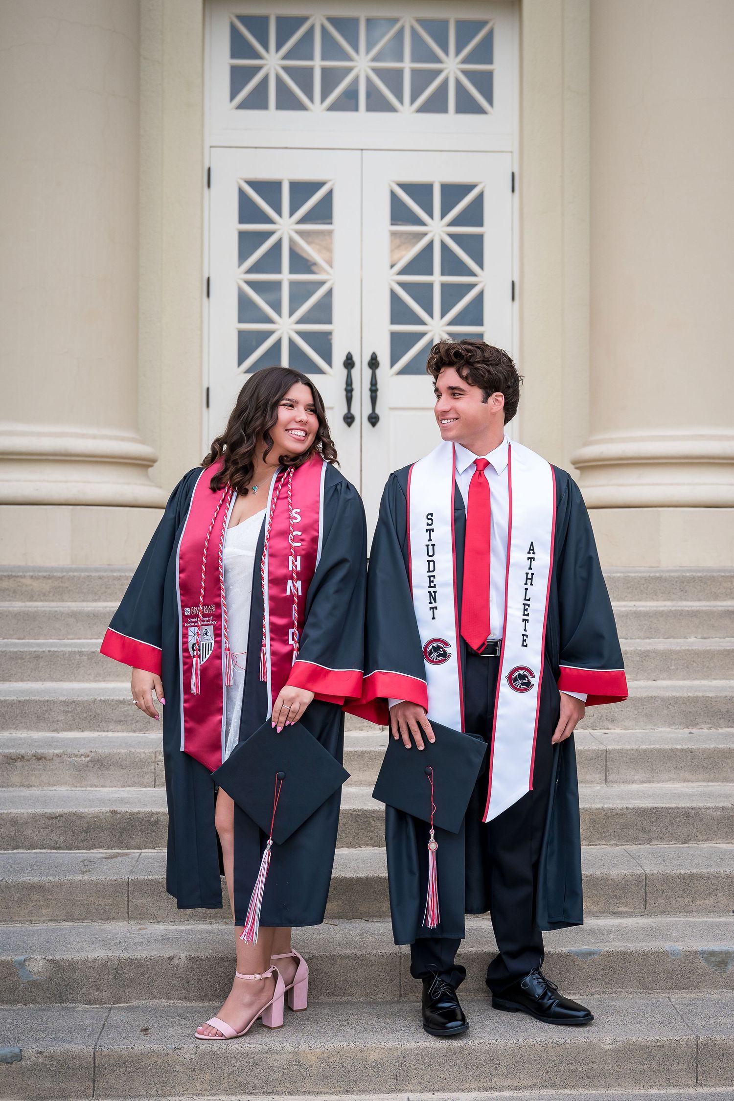 Twin graduates standing side by side in front of Memorial Hall at Chapman University