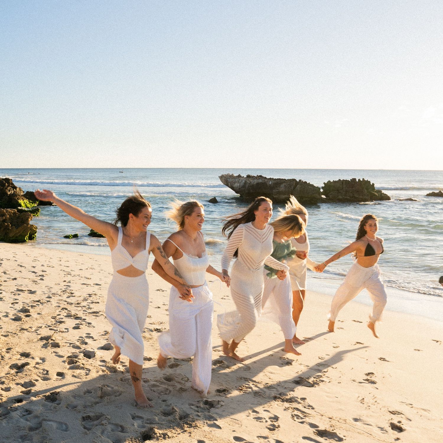 A group in white outfits dances joyfully along a scenic beach with rocky outcrops and ocean waves in the background.