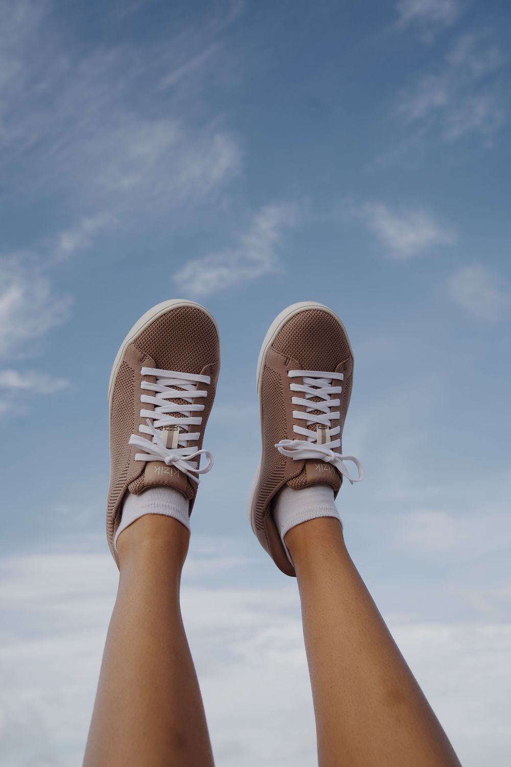 Brown canvas sneakers photographed against blue sky background with feet pointed upward.