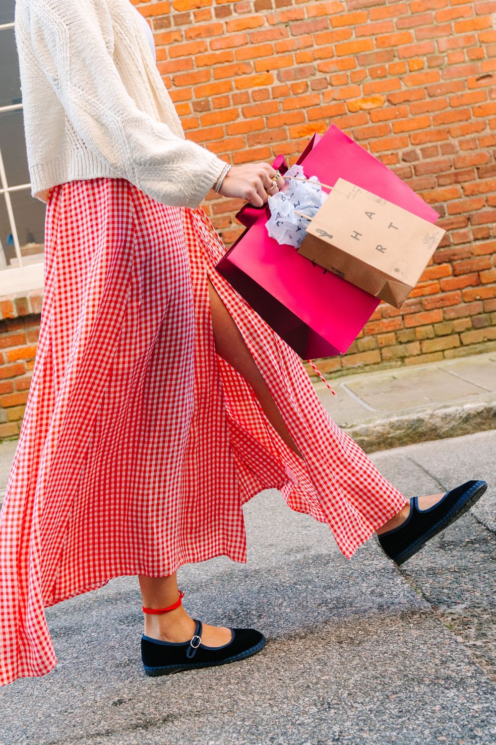 A person wearing a red gingham skirt and cream sweater walks on pavement carrying pink shopping bags.