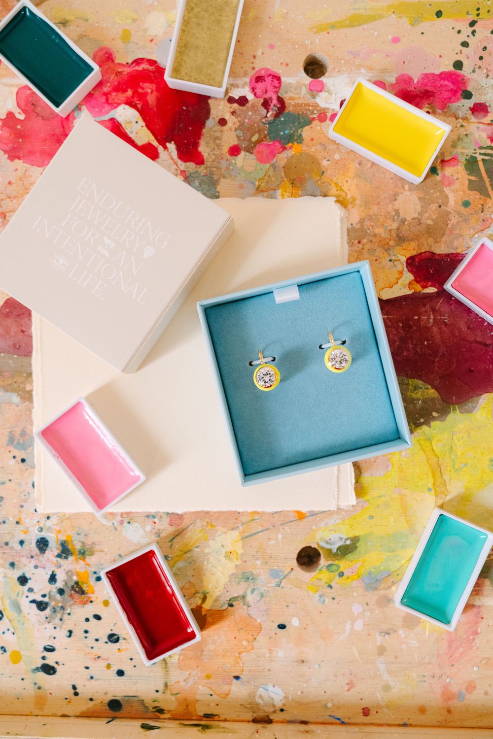 Colorful paint splatters and geometric paper shapes create an abstract artistic composition on a textured surface.