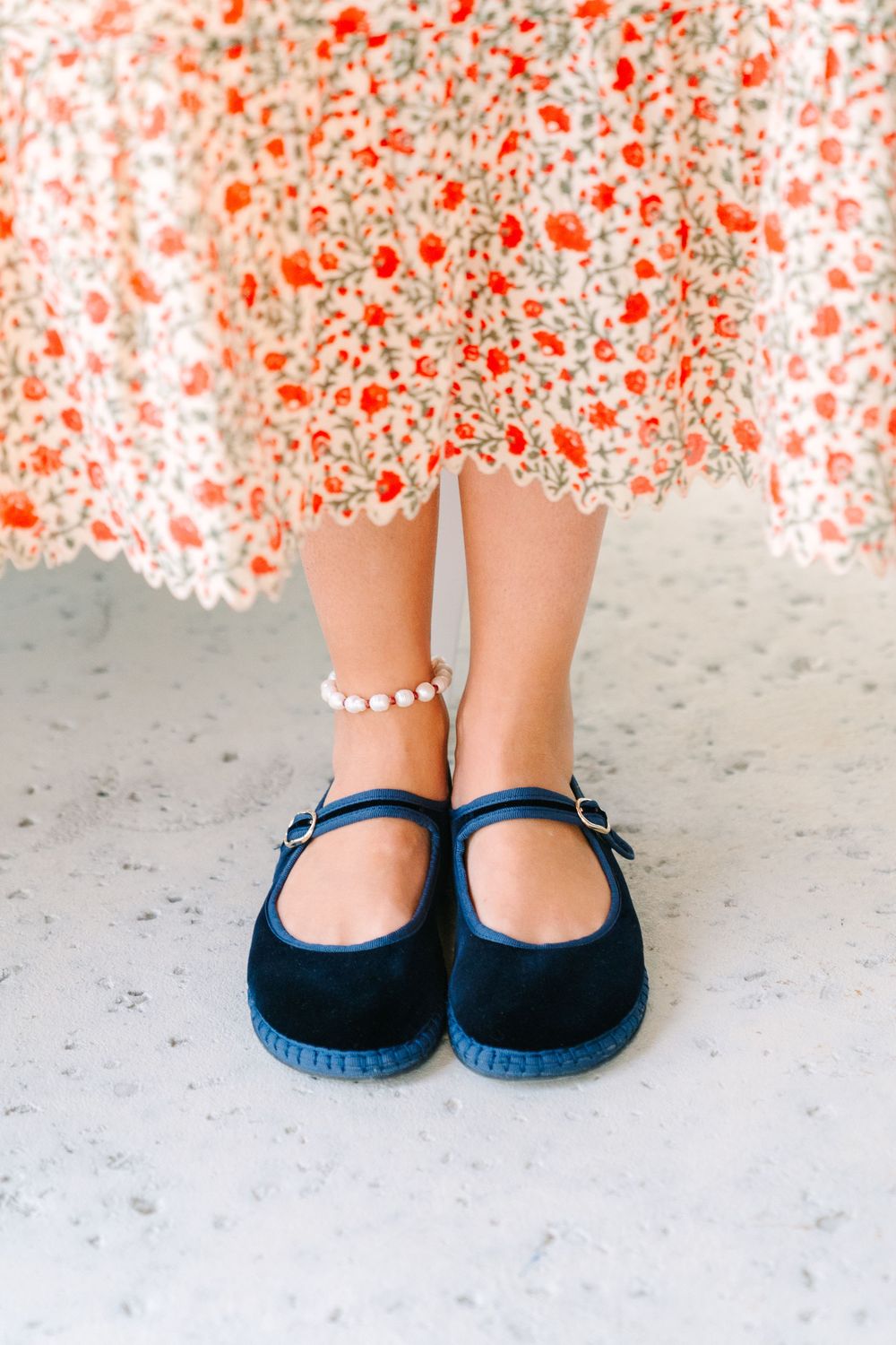 A pair of navy blue Mary Jane shoes with white ankle socks against a light colored floor, with a floral dress visible.