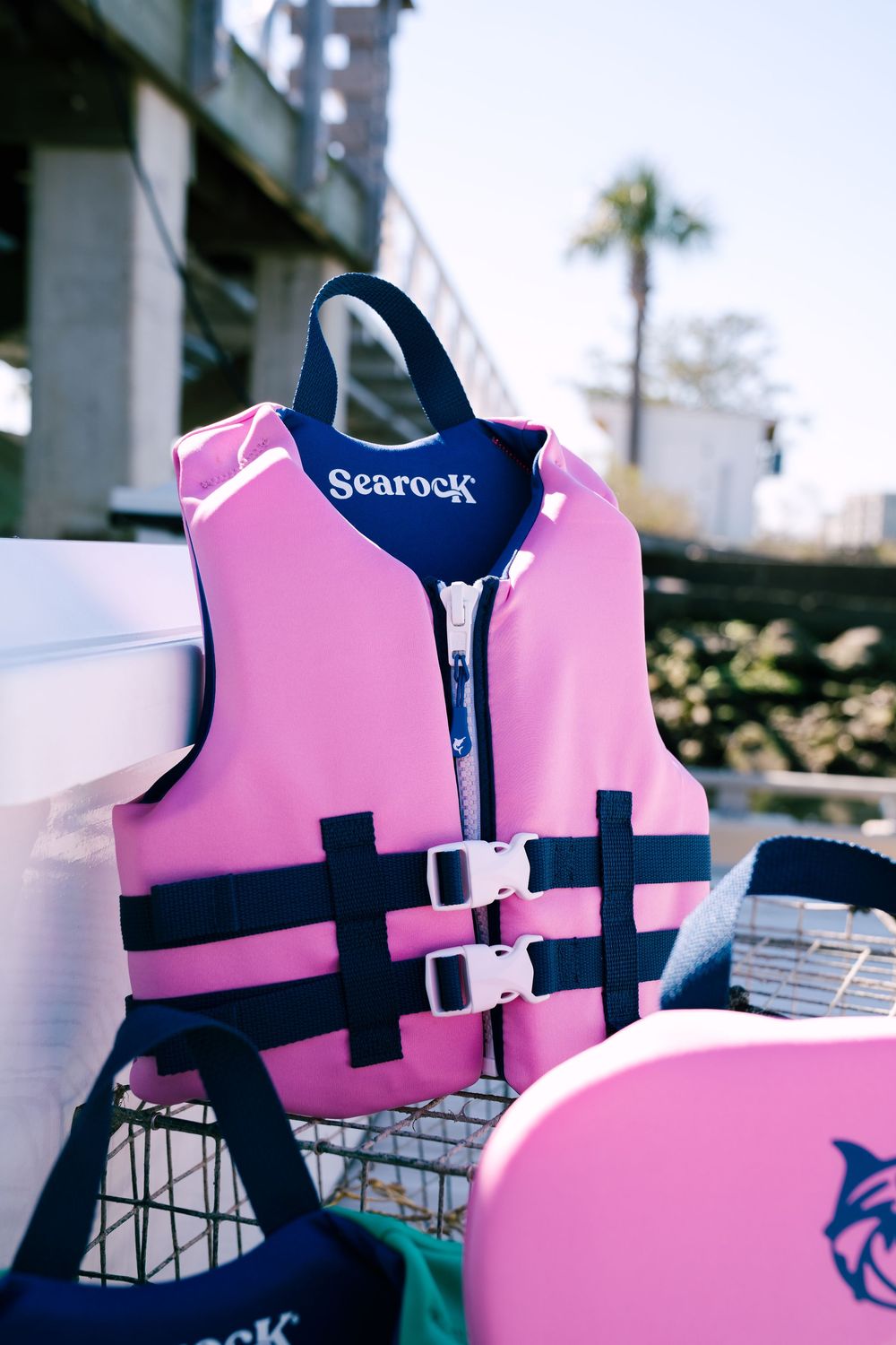 Pink and navy Searock life vest with buckle straps sits on display with palm trees in background.