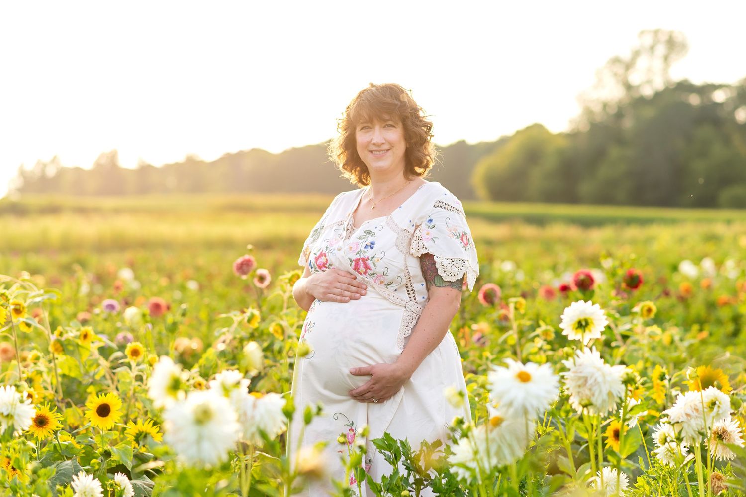 An expectant mother in a white dress stands among vibrant sunflowers at sunset at the Flower Lady and Gent.