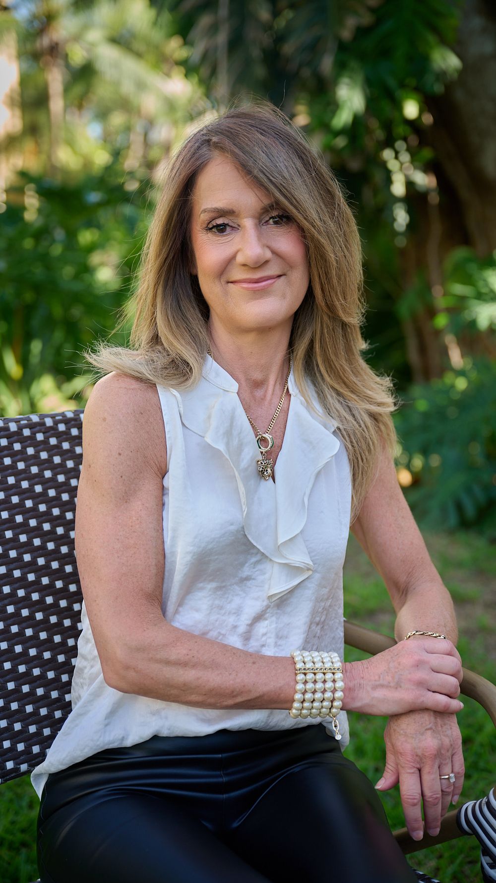 A woman in a white ruffled blouse and dark skirt sits casually on outdoor patio furniture in a garden setting.