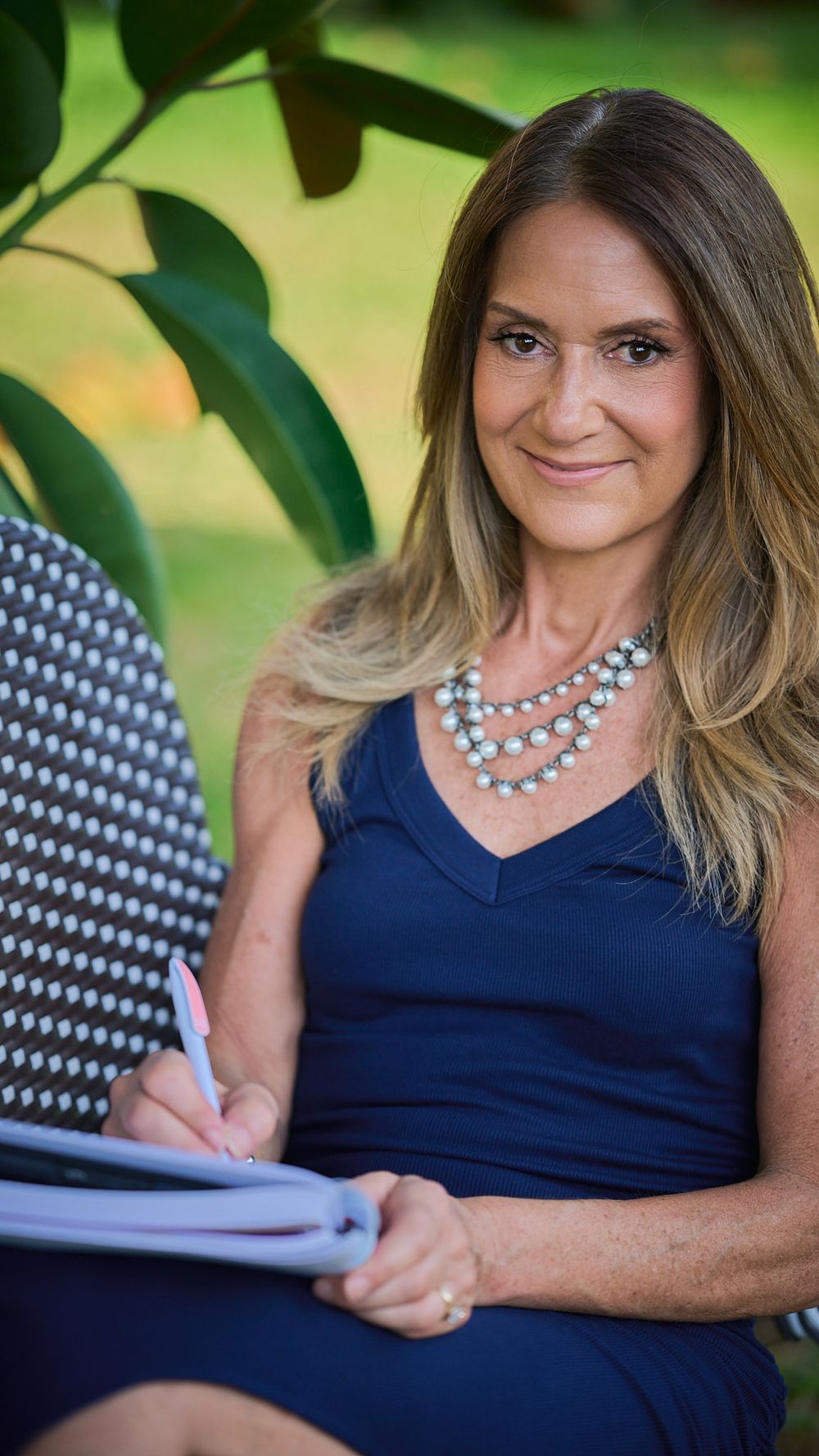 Professional woman in navy dress with pearl necklace writes in notebook while seated outdoors near green foliage.