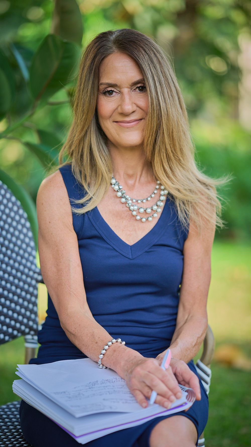 Woman in navy blue dress and pearl necklace sits outdoors writing in notebook while smiling at camera.
