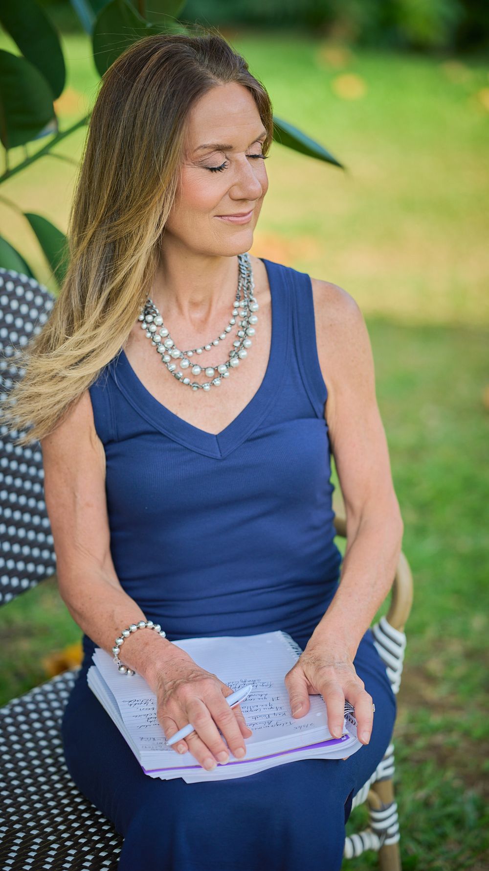 Woman in blue sleeveless dress writes in notebook while sitting outdoors in garden setting.
