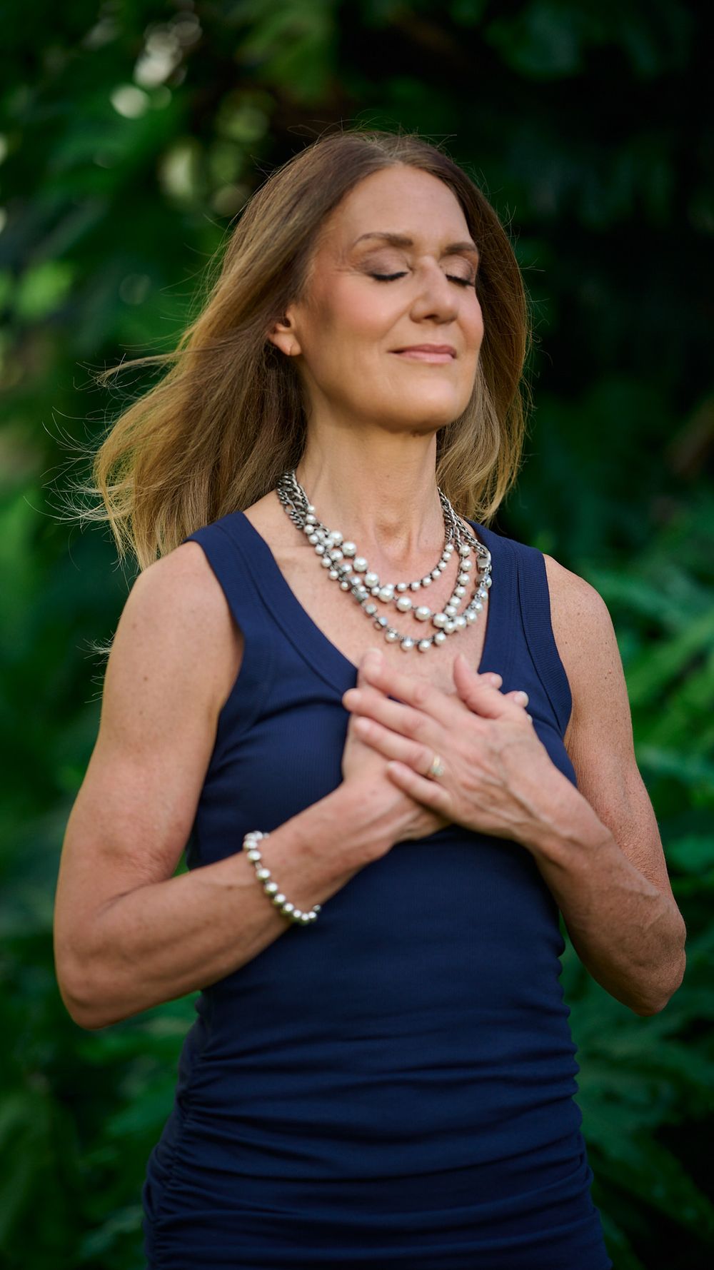 Woman in navy sleeveless top and pearl necklace strikes series of graceful emotional poses against green foliage background.