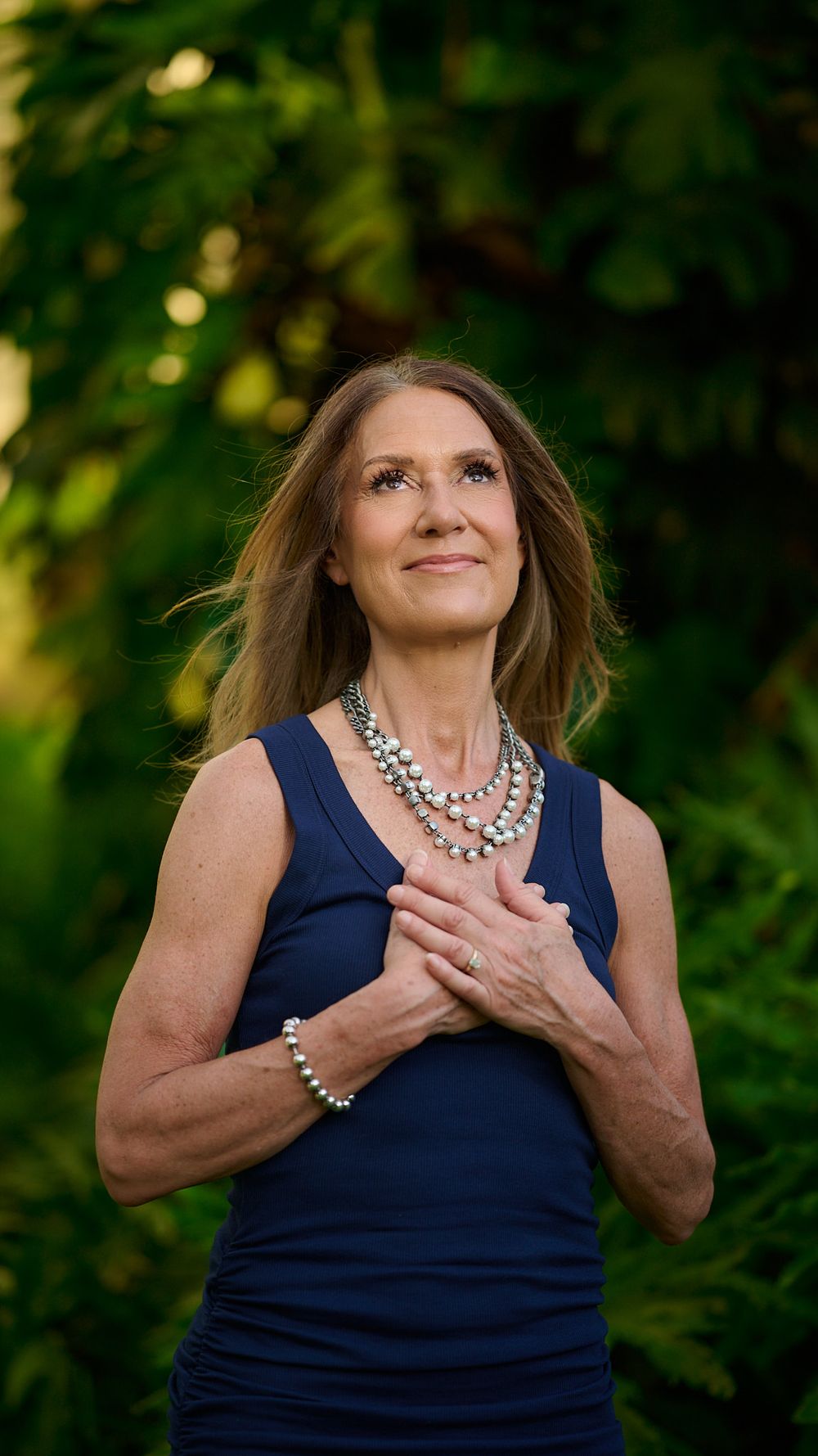 Portrait of person in navy sleeveless dress with layered pearl necklaces looking up against green foliage background.