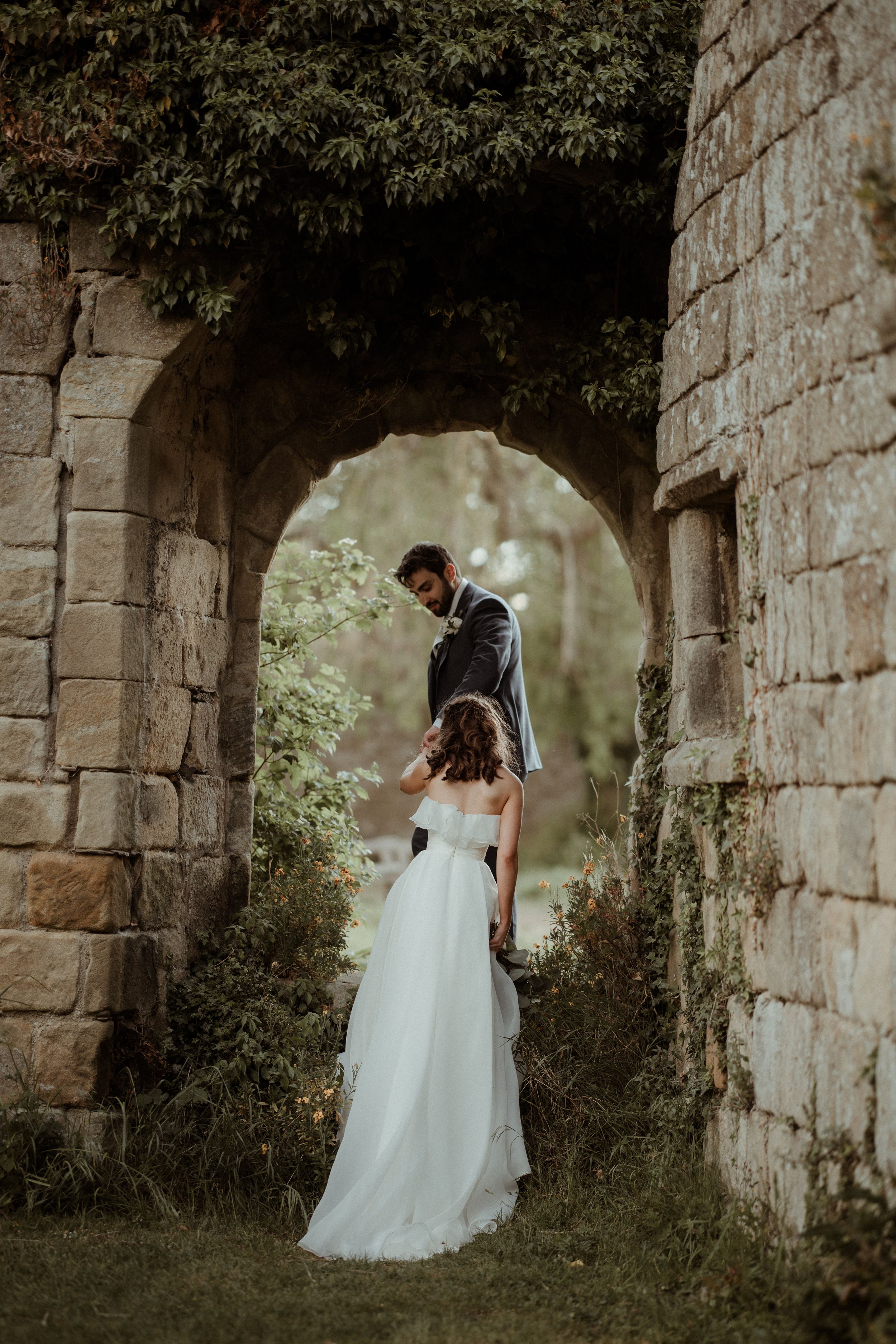 A couple in formal wedding attire pose romantically under an old stone archway surrounded by greenery.