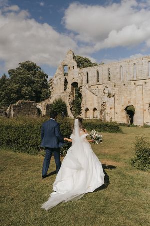 A couple in wedding attire stand on the lawn in front of an ancient stone abbey ruin under a cloudy summer sky.
