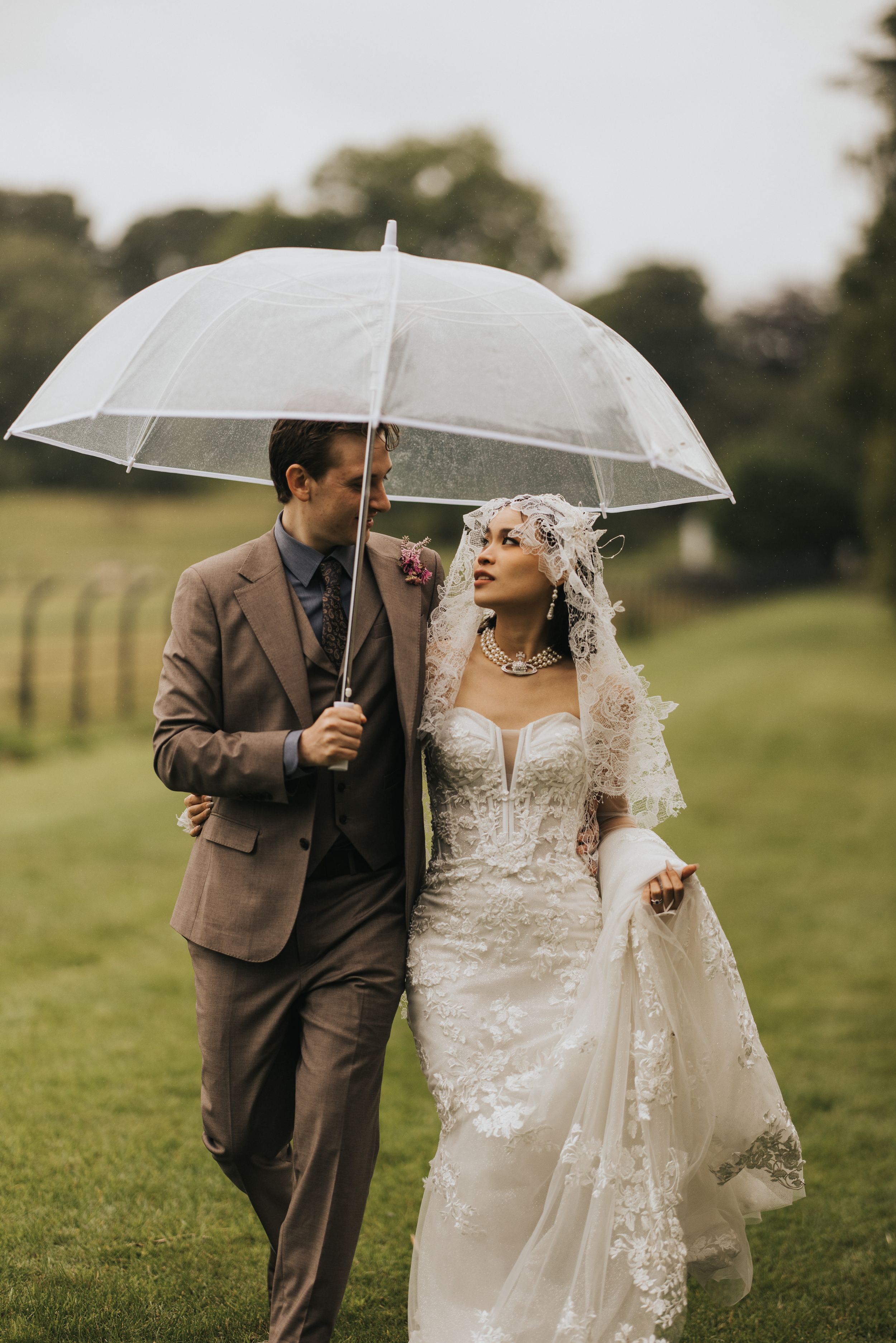 Couple shares clear umbrella on rainy wedding day in countryside with lush green field and trees in background.