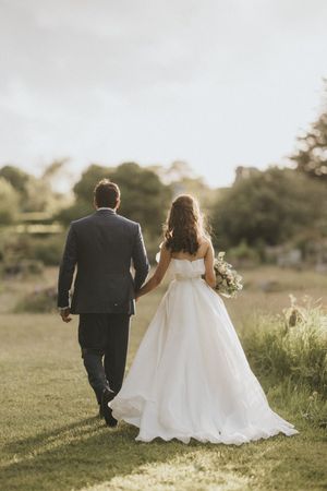 A couple in wedding attire walk hand in hand through a sunlit meadow at golden hour in their white gown and dark suit.