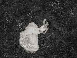 A person in a flowing white dress lies gracefully in a dark meadow, captured in black and white from above.