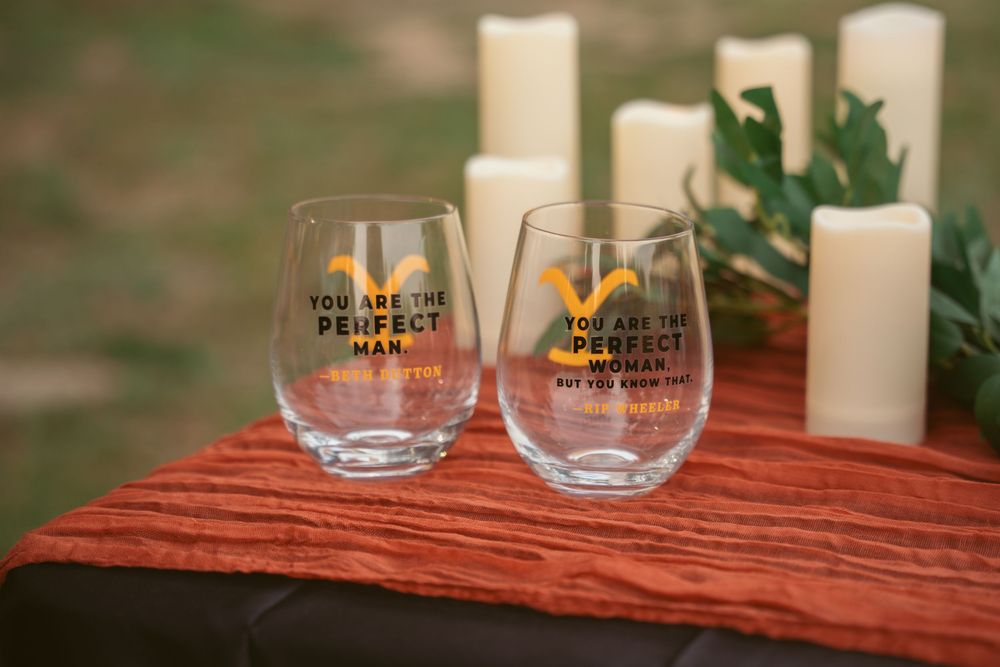 Two stemless wine glasses with text printed on them sit on a red tablecloth next to white candles.