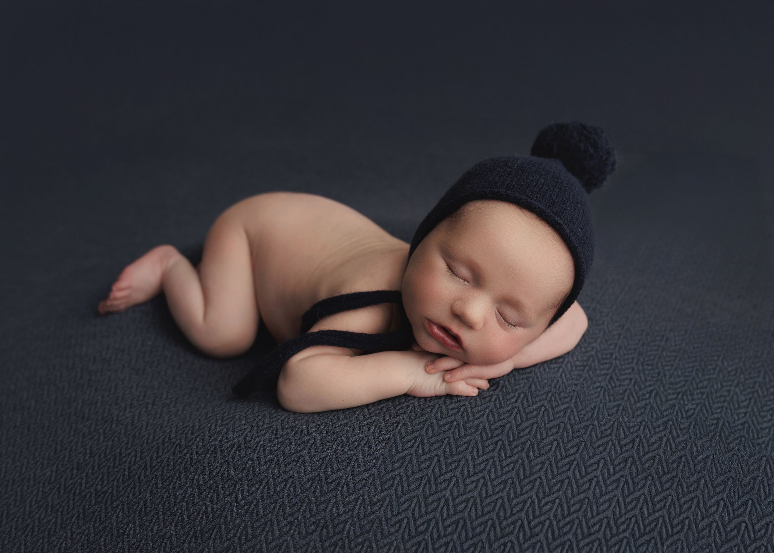 A sleeping newborn baby lies peacefully on a dark textured background in a minimalist portrait style.