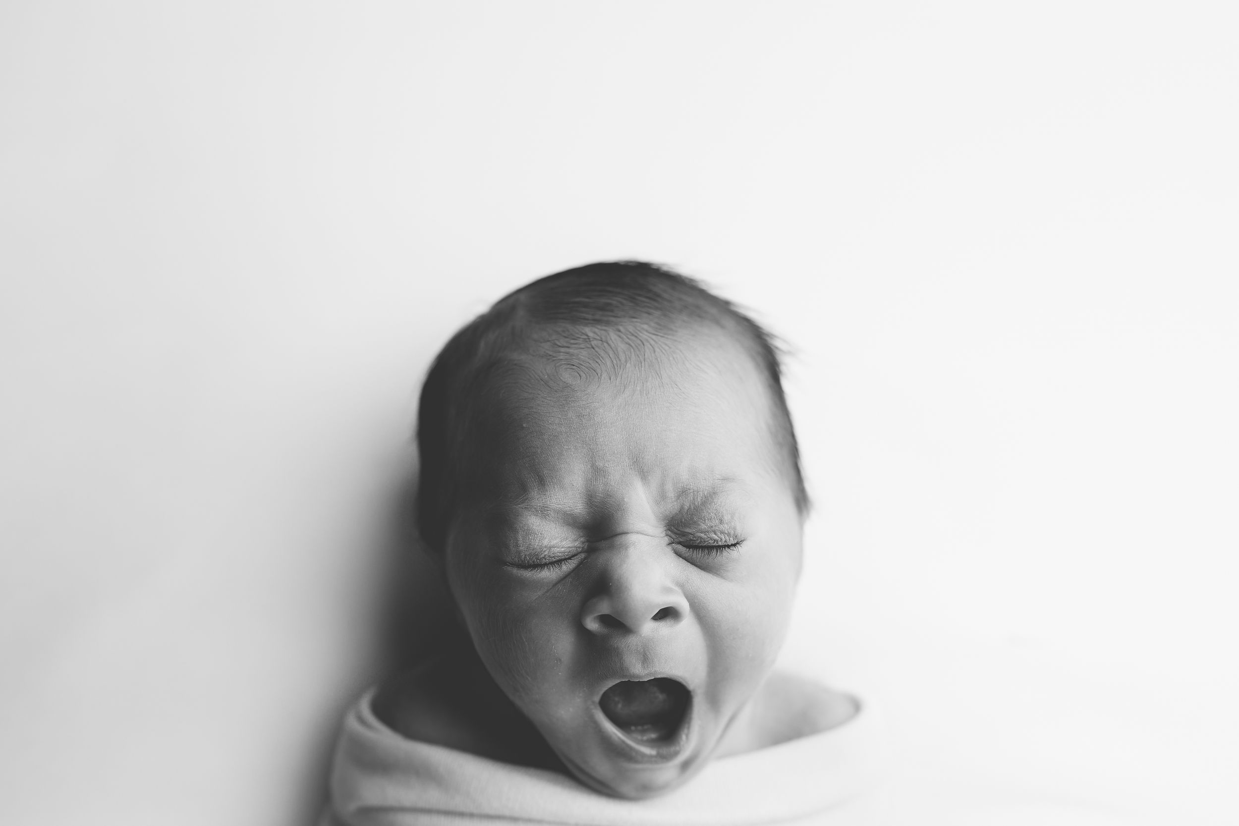 Black and white portrait of a yawning newborn baby wrapped in white cloth against light background.