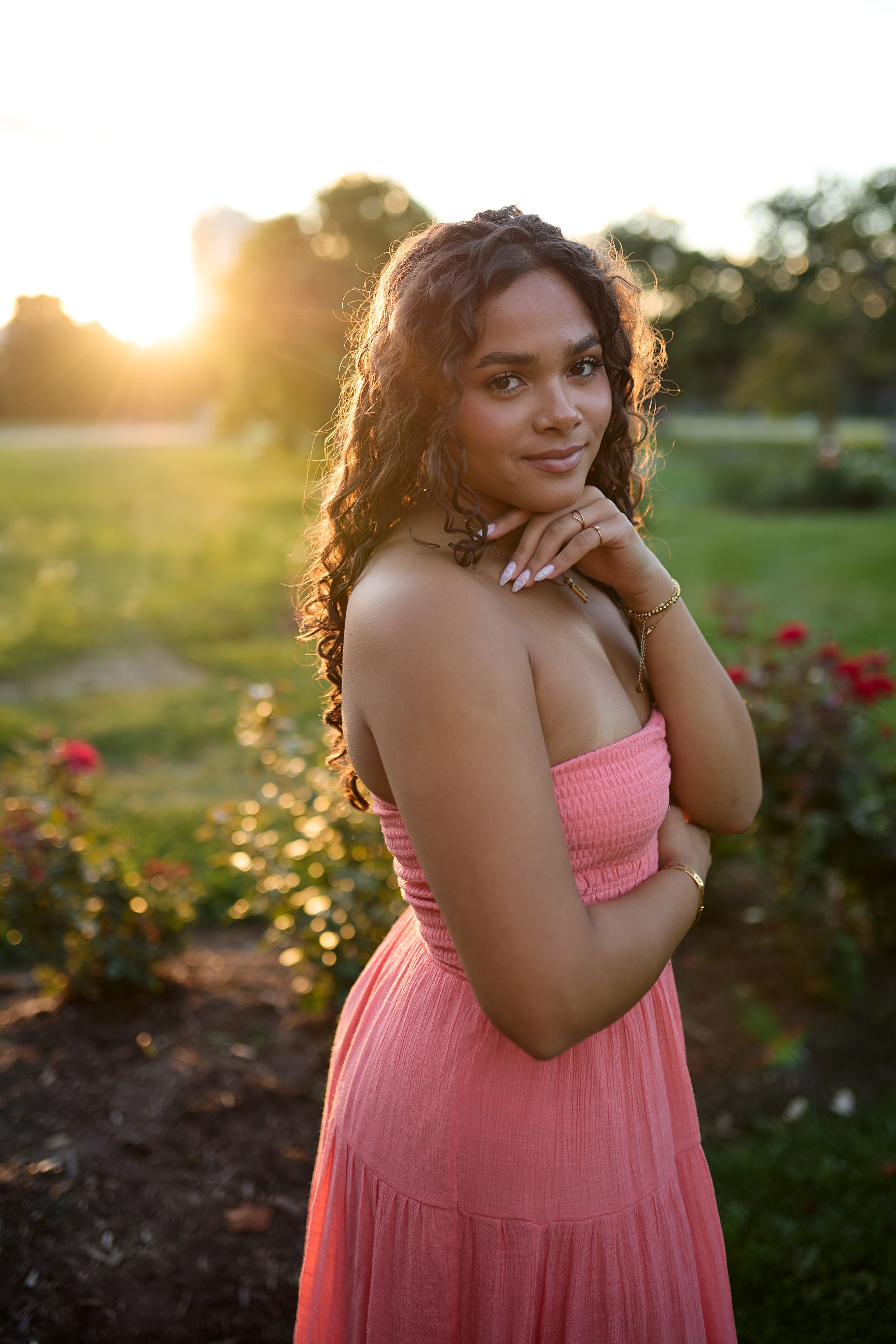 A person in a pink sundress poses in a garden at sunset among blooming roses.