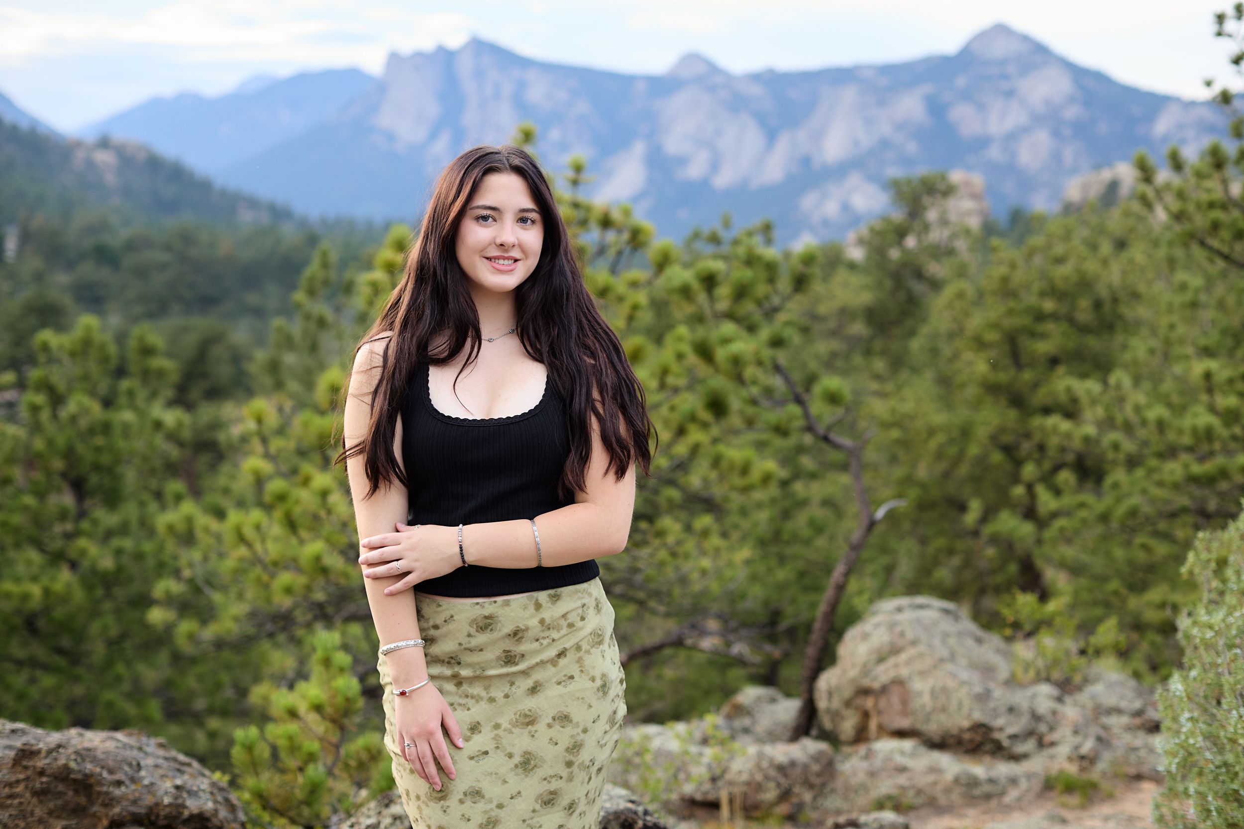 Woman poses on large rocks with scenic mountain views and green forest landscape in the background.