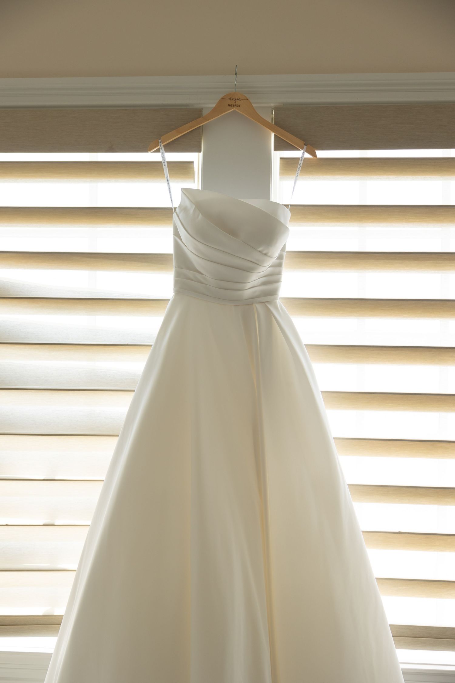 An elegant white wedding dress with a ruched bodice hangs from a wooden hanger against illuminated window blinds.