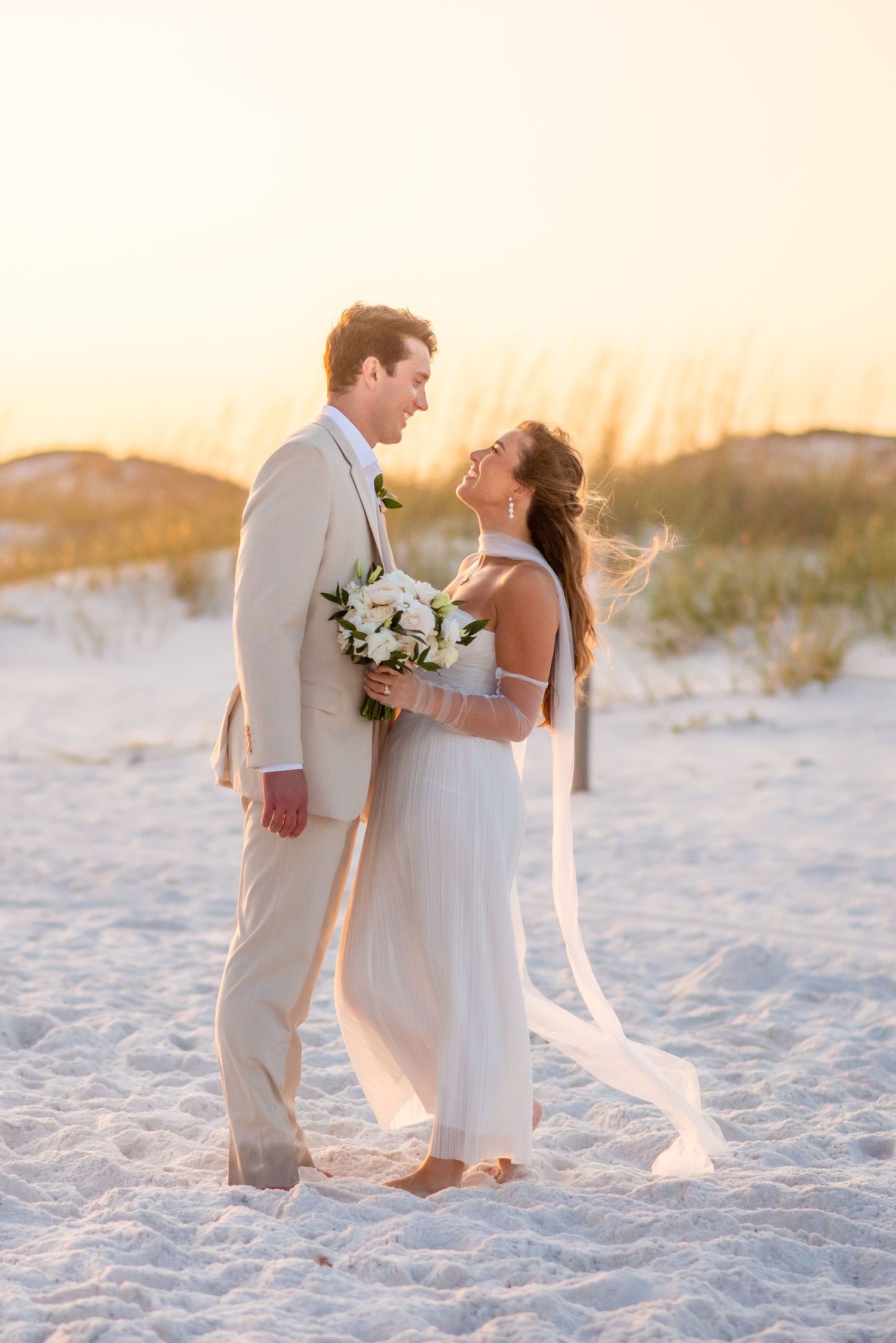 A couple embraces on a beach at sunset, their white outfits glowing against the golden light and ocean waves.