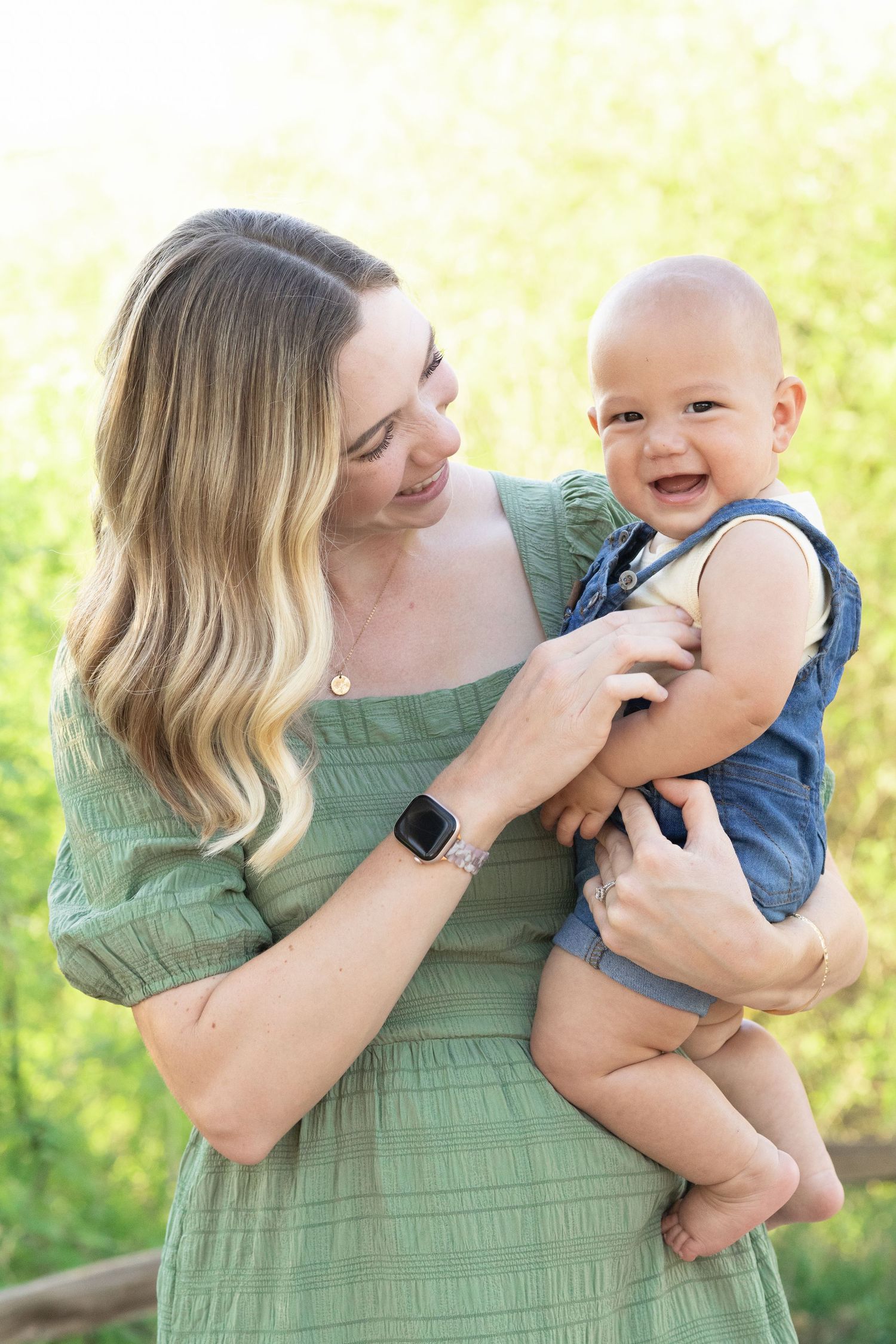 Mom tickling infant son in front of tall green bushes and grass at the Gilbert Riparian Preserve.