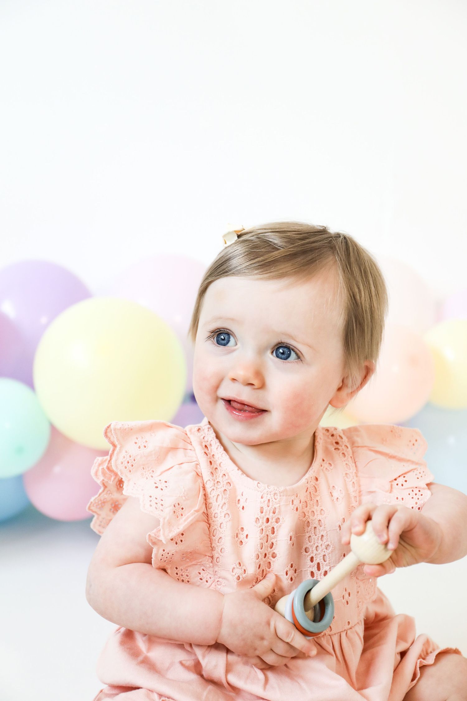 Sweet baby in peach dress poses with soft pastel balloons during gentle and dreamy photography session, during birthday shoot in Nottingham