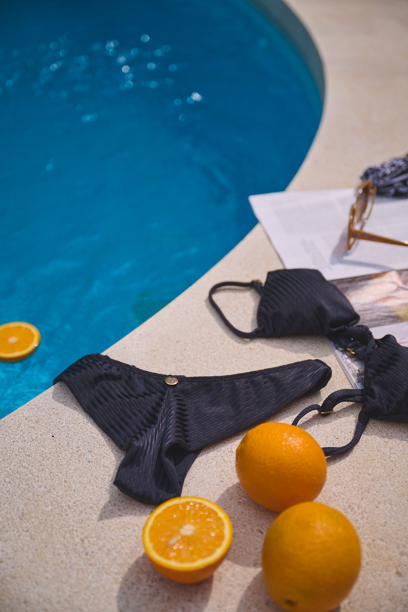 Black bikini and face mask laid by pool edge with fresh oranges scattered around blue swimming pool water.