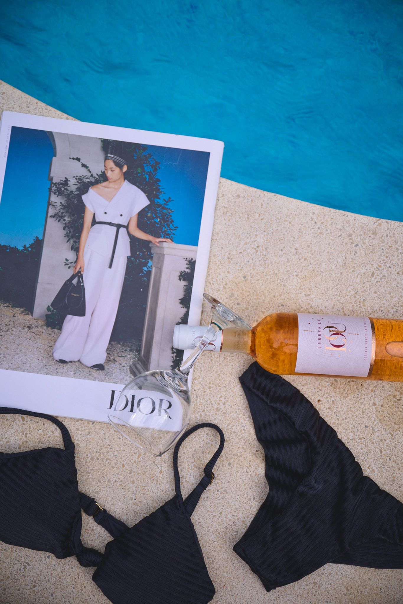 A poolside flatlay with fashion items including a Dior photo, black swimwear pieces, and a sunscreen bottle.