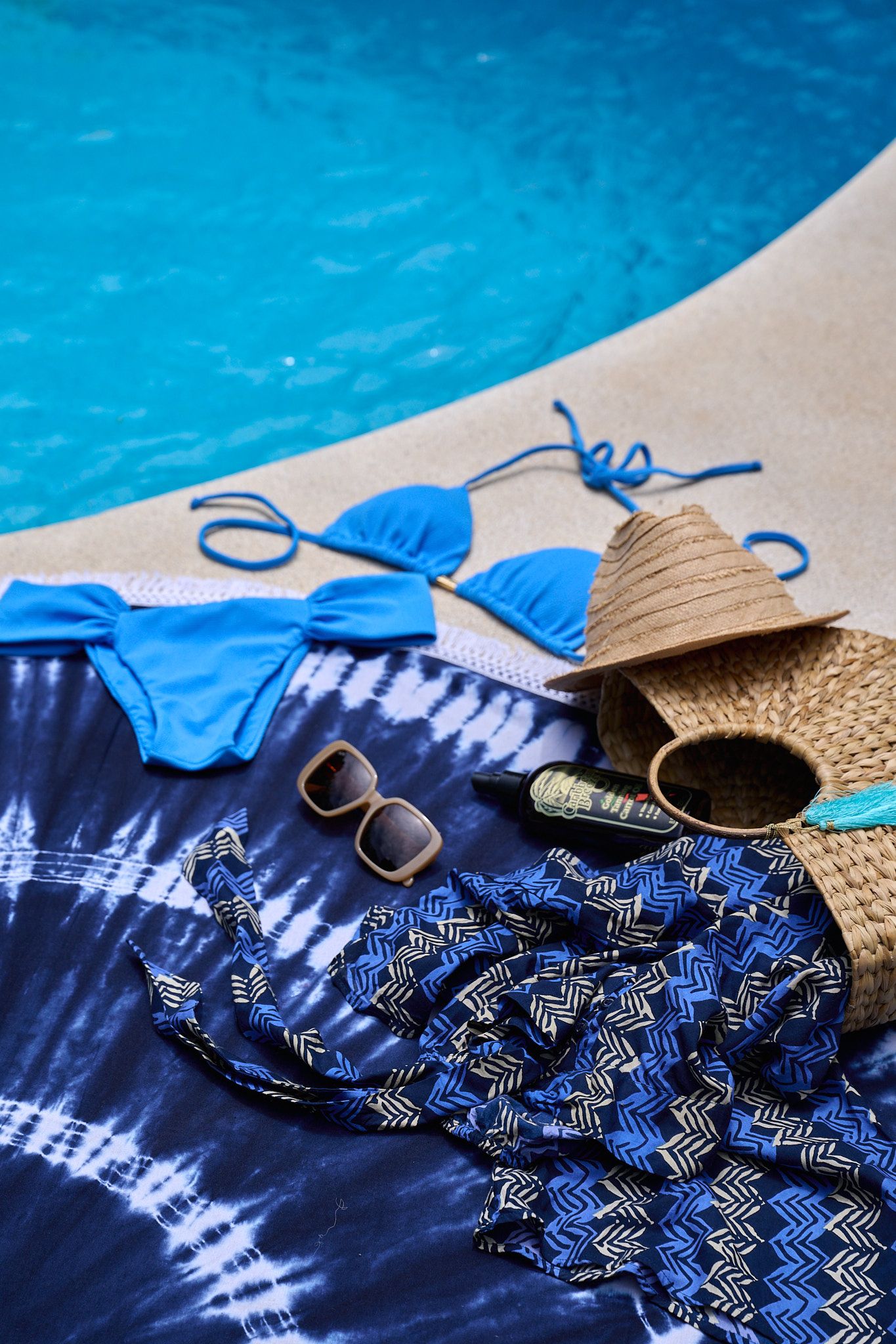 Summer poolside essentials including blue bikini, straw hat, sunglasses, and navy tie-dye beach dress by swimming pool.