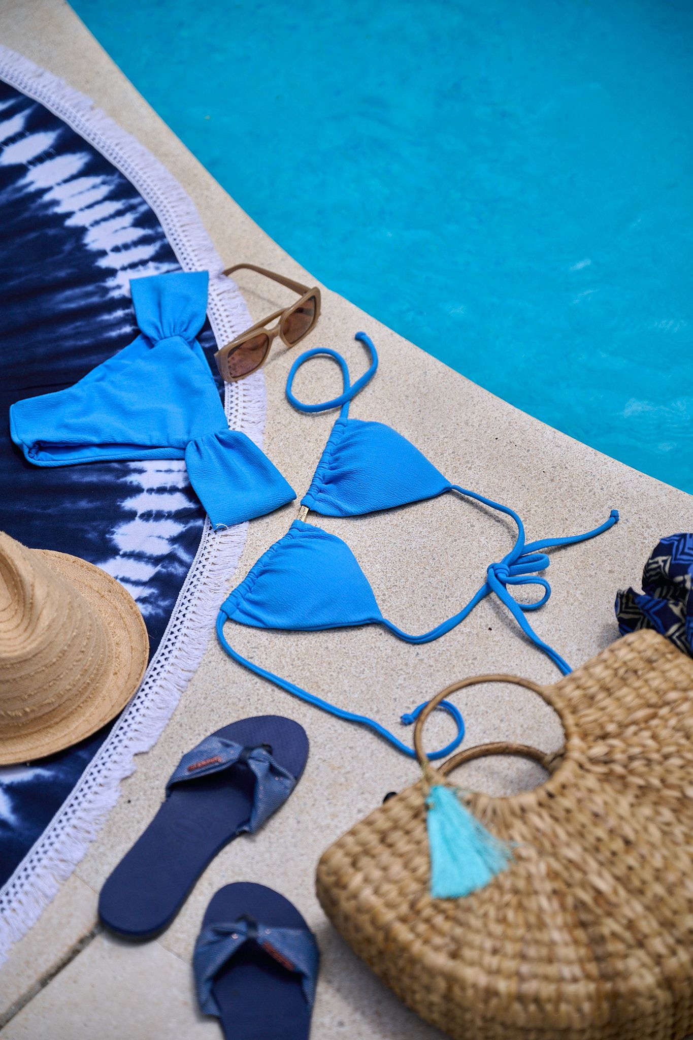 Beach items including a straw hat, blue swimsuit, and flip flops arranged by a pool's edge.