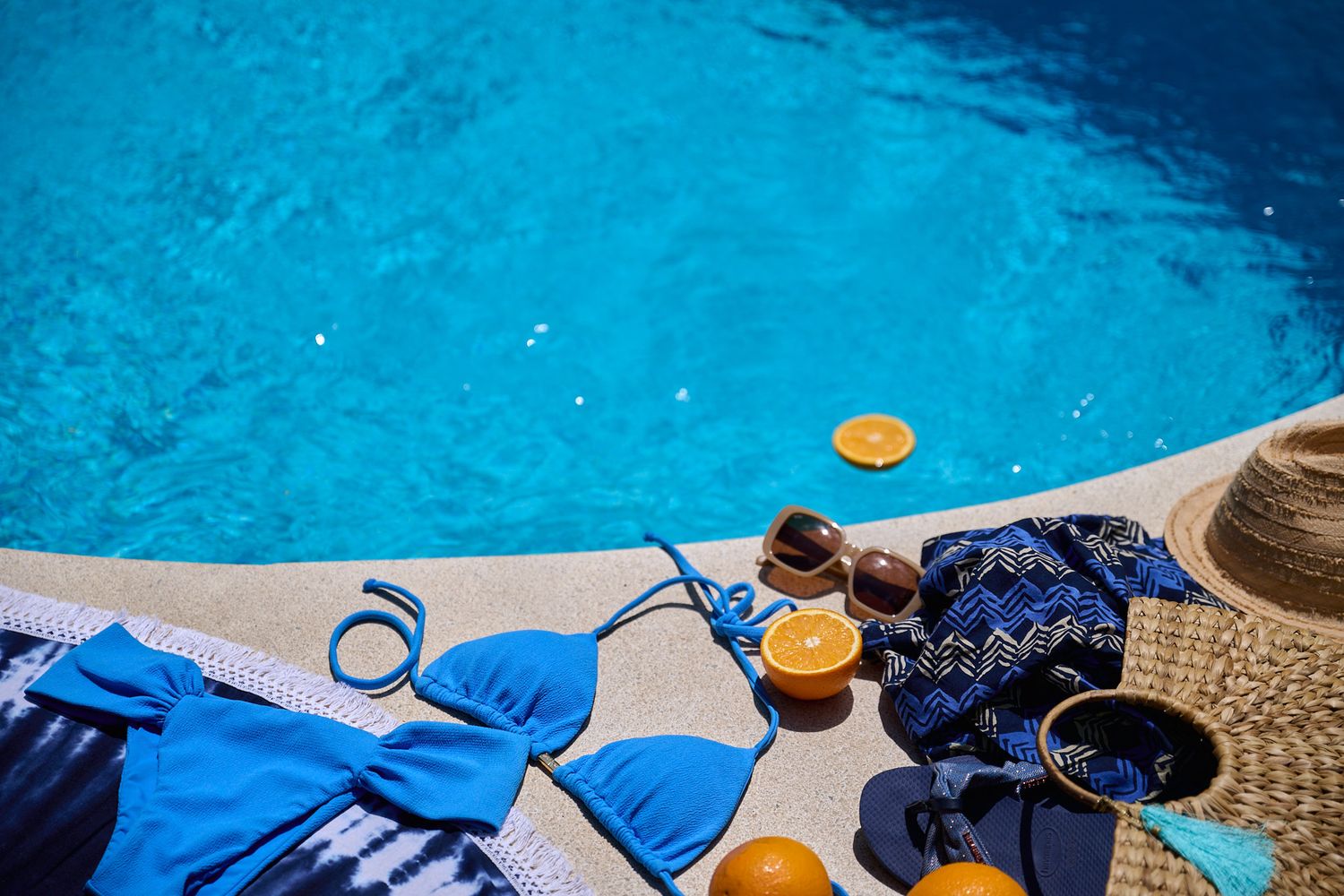 A poolside scene with vibrant blue swimwear, fresh oranges, and a straw hat against crystal clear turquoise water.