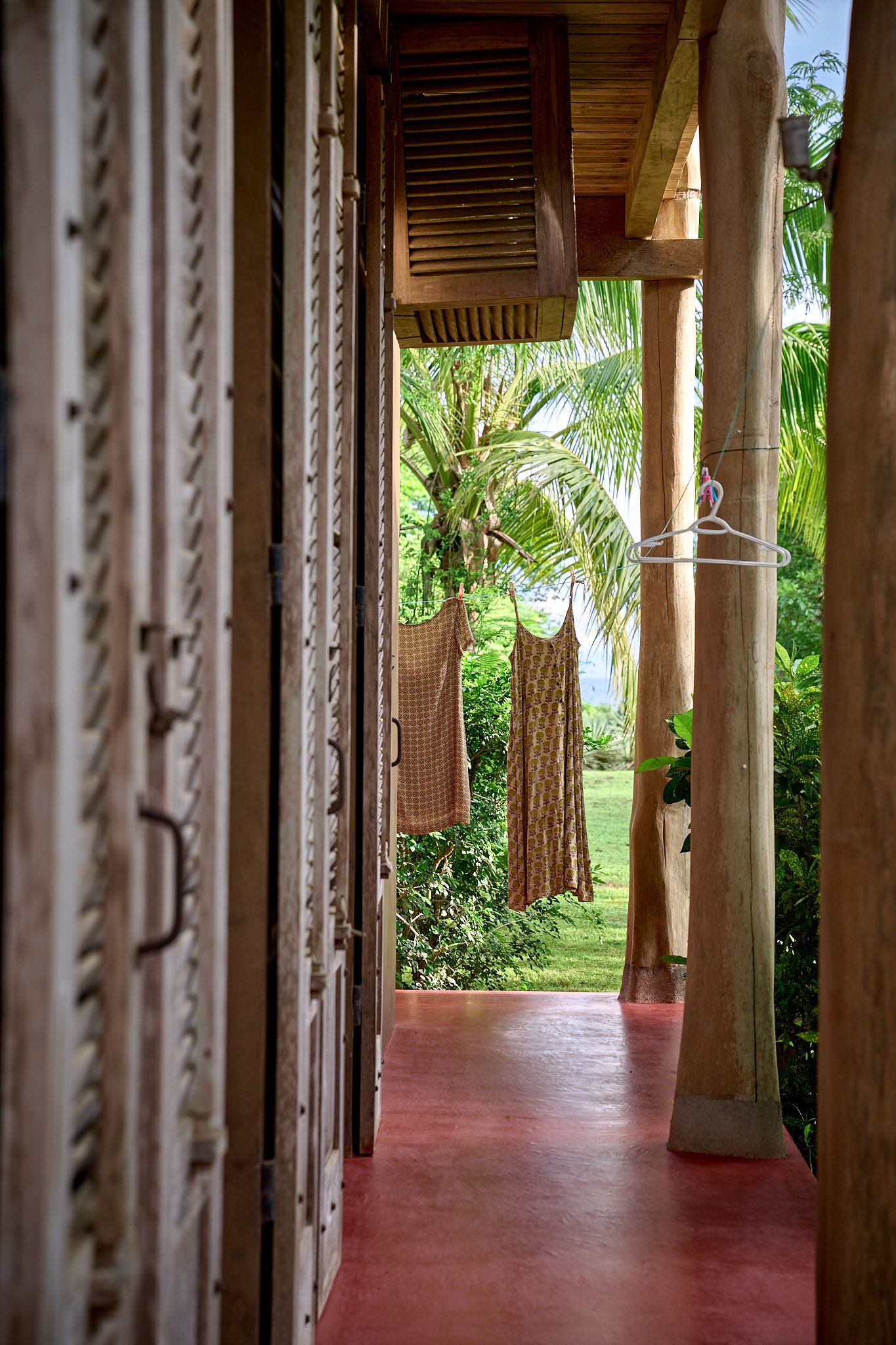 A covered porch with wooden shutters and columns leads to a tropical garden view with palm trees and lush greenery.