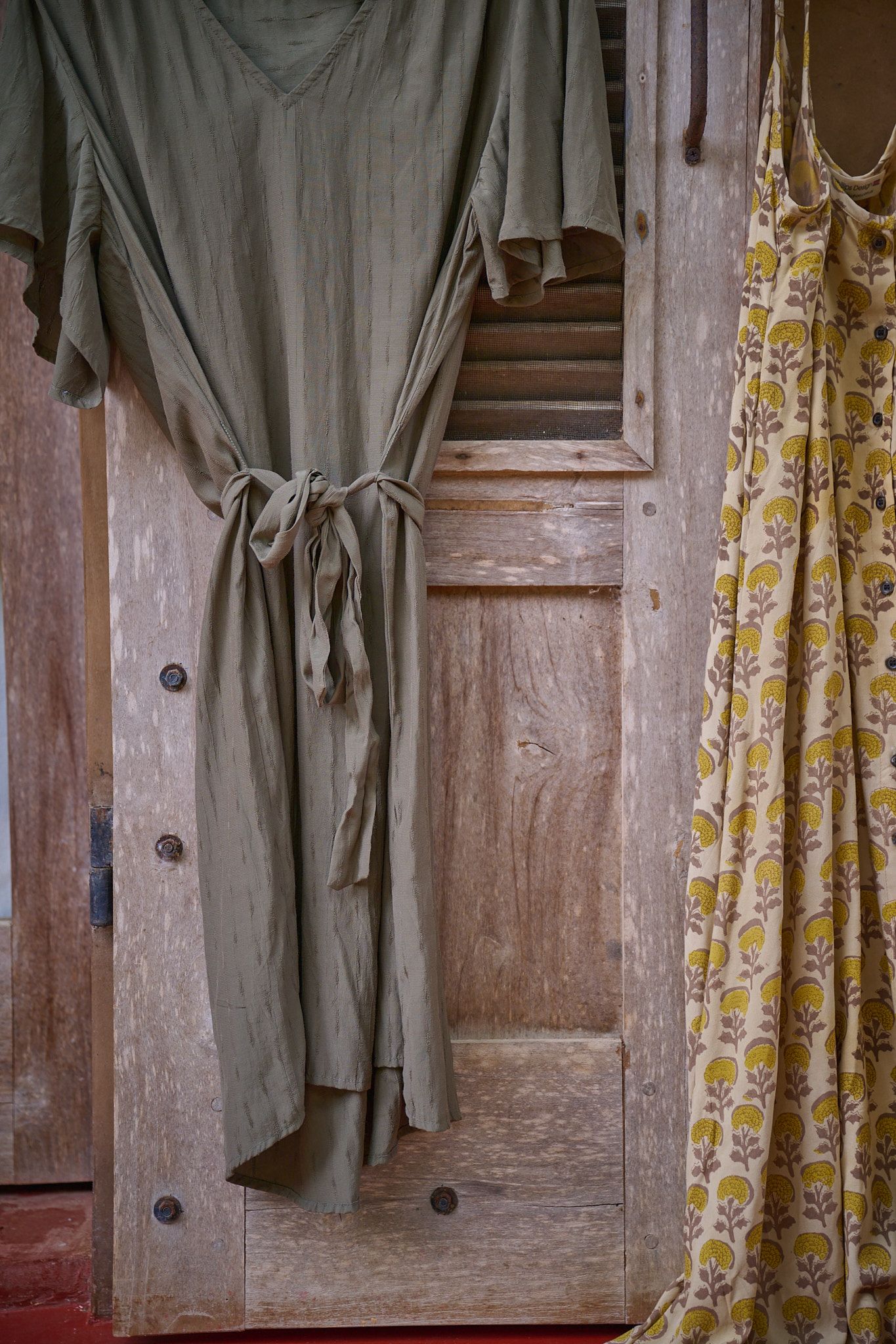 A sage green dress hangs on a wooden door alongside a yellow patterned curtain in a vintage-style setting.