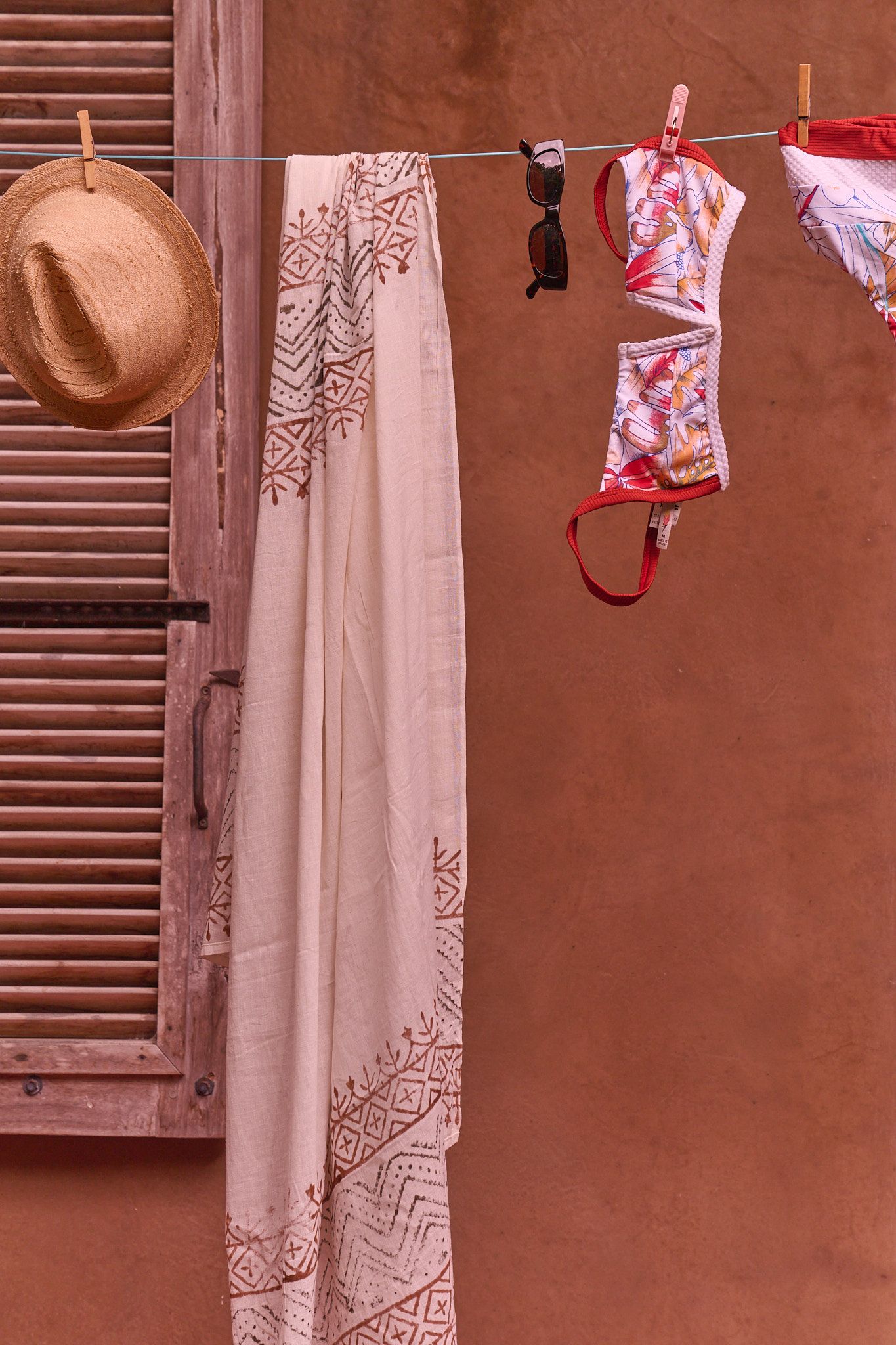 A straw hat and patterned clothes hang on a clothesline against a terracotta wall with shuttered window.