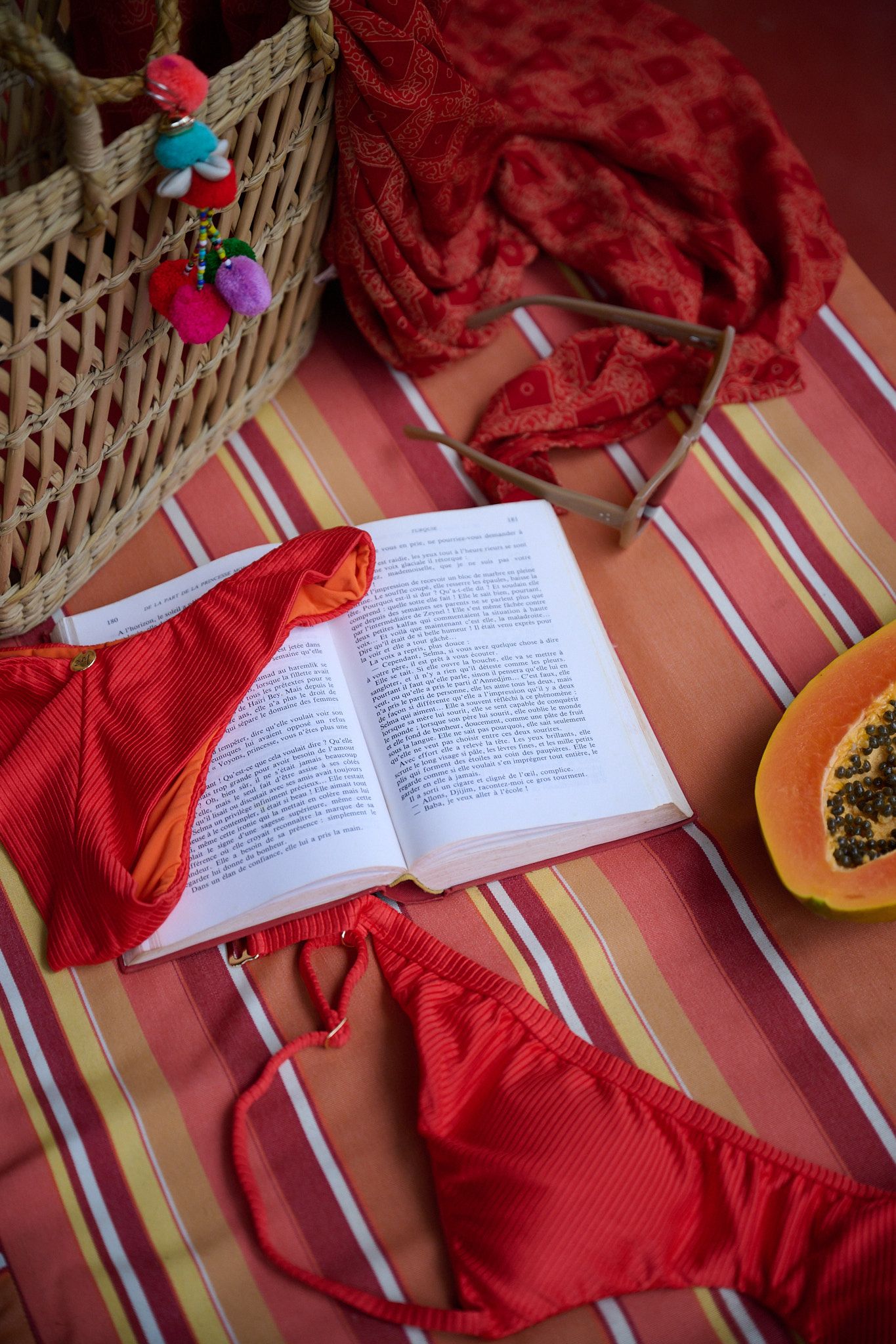 An open book lies on a striped surface with red fabric and a wicker basket arranged around it.