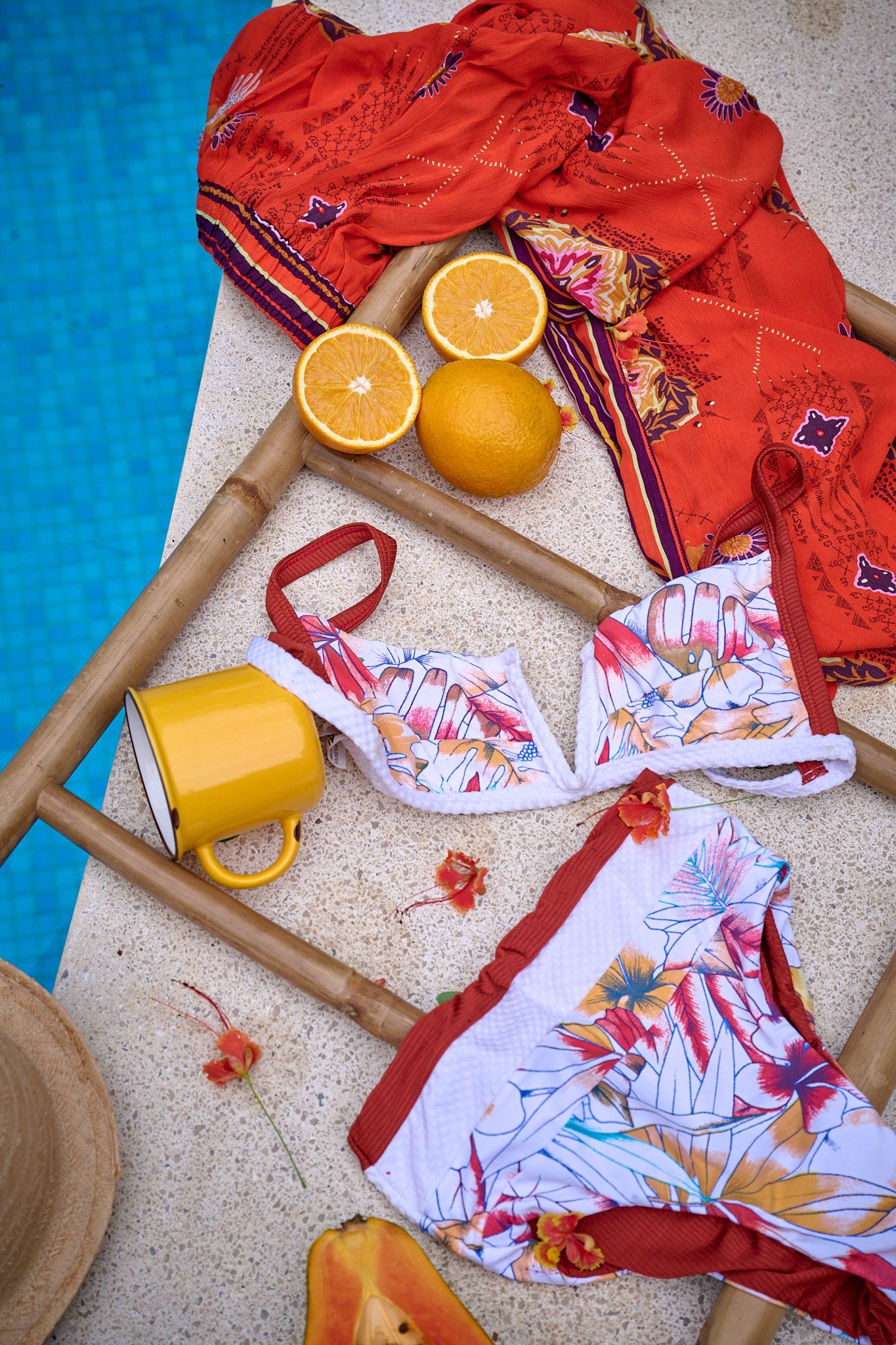 Flatlay of bright orange citrus fruits with red patterned fabric and white clothing items on a textured beige surface.