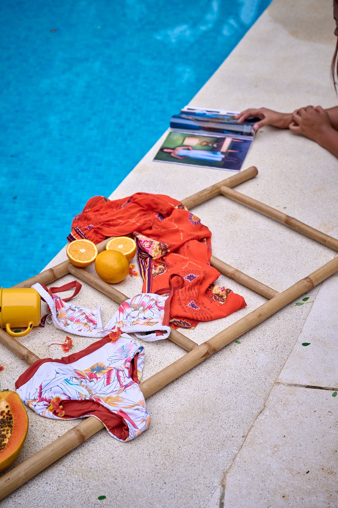 Colorful swimwear and fresh oranges laid out on wooden steps beside a bright blue swimming pool.