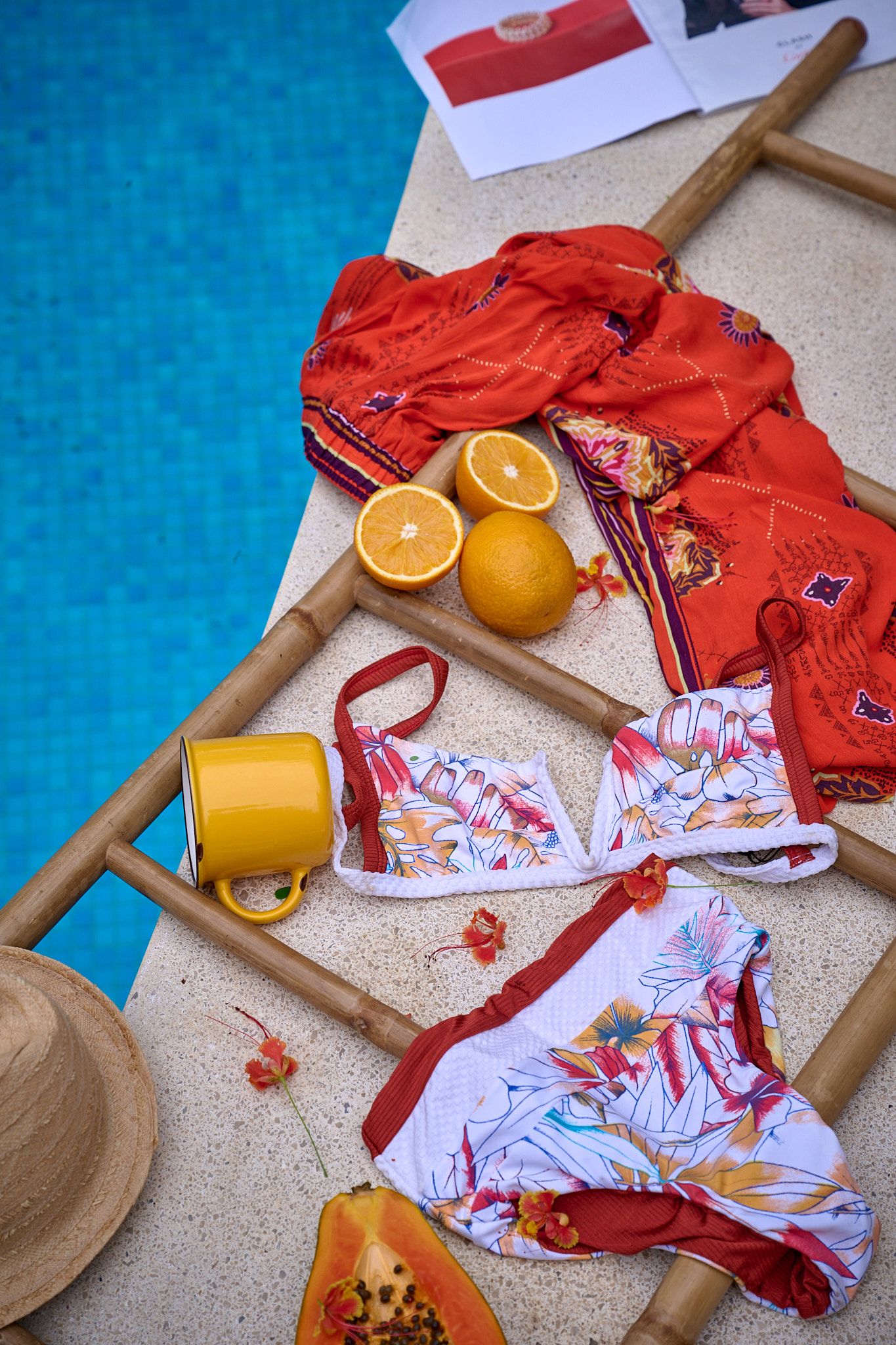 Bright red and white patterned swimsuits with oranges scattered on a wooden hanger against a vibrant blue background.