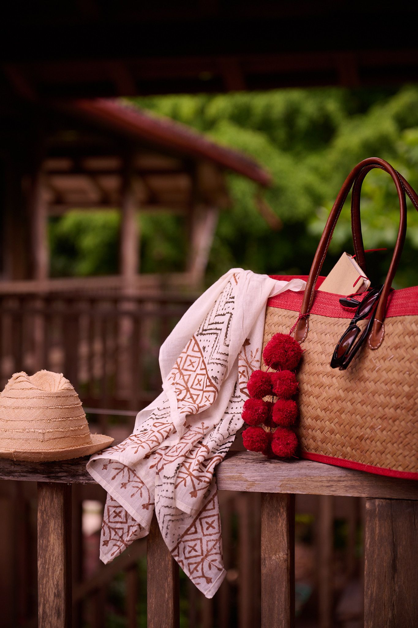 A woven straw bag with red embroidered cloth and pom poms sits on a wooden railing against a blurred green background.
