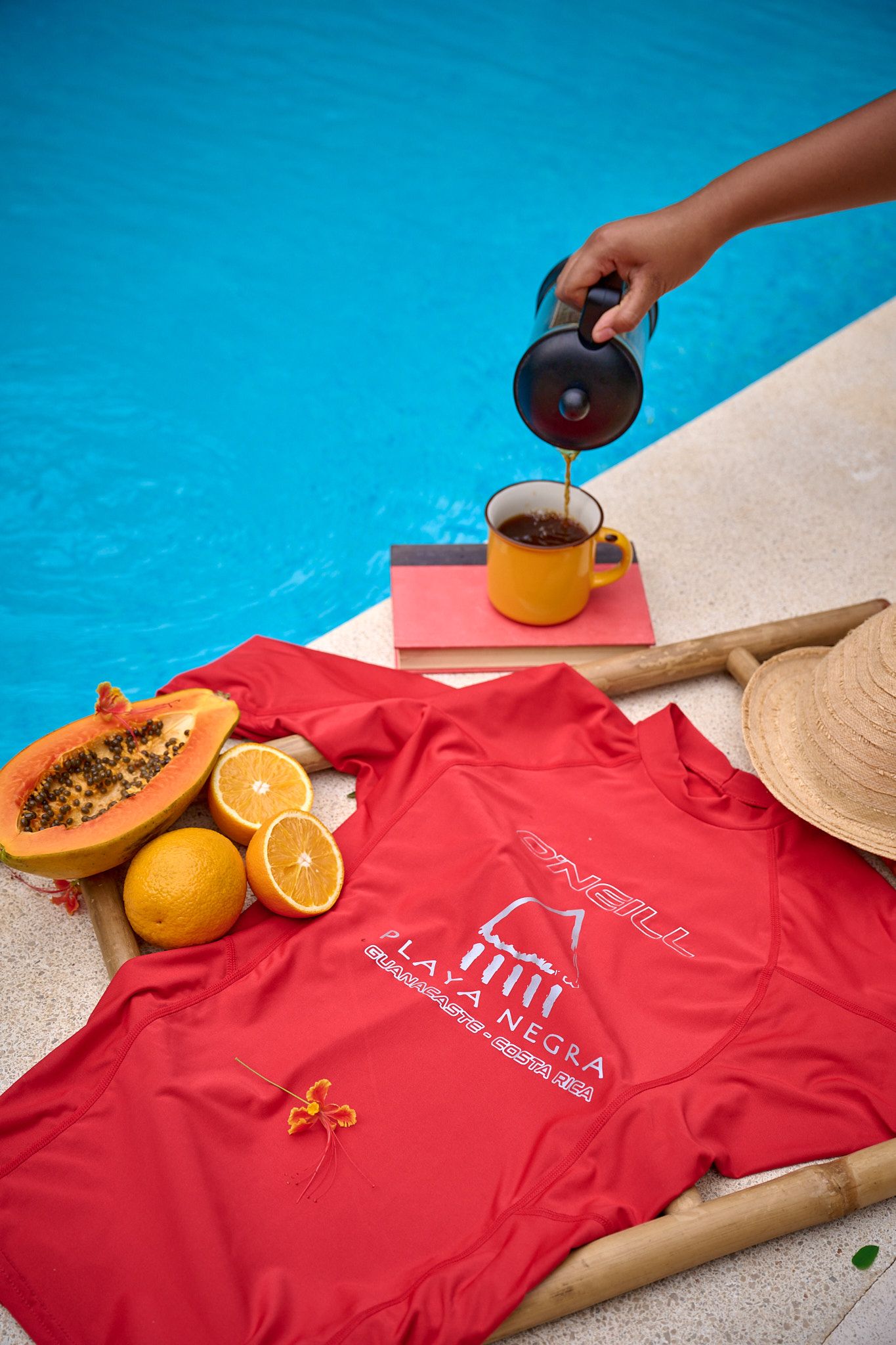 Coffee being poured by pool with red t-shirt, tropical fruits and straw hat on sunny day.