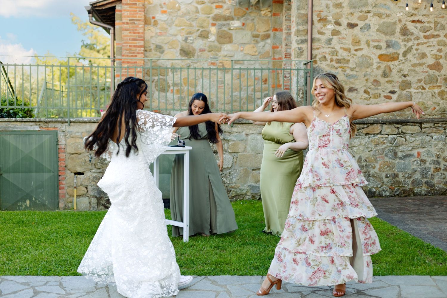Three bridesmaids in flowing dresses dance and spin playfully on the grass near a stone wall during an outdoor wedding.