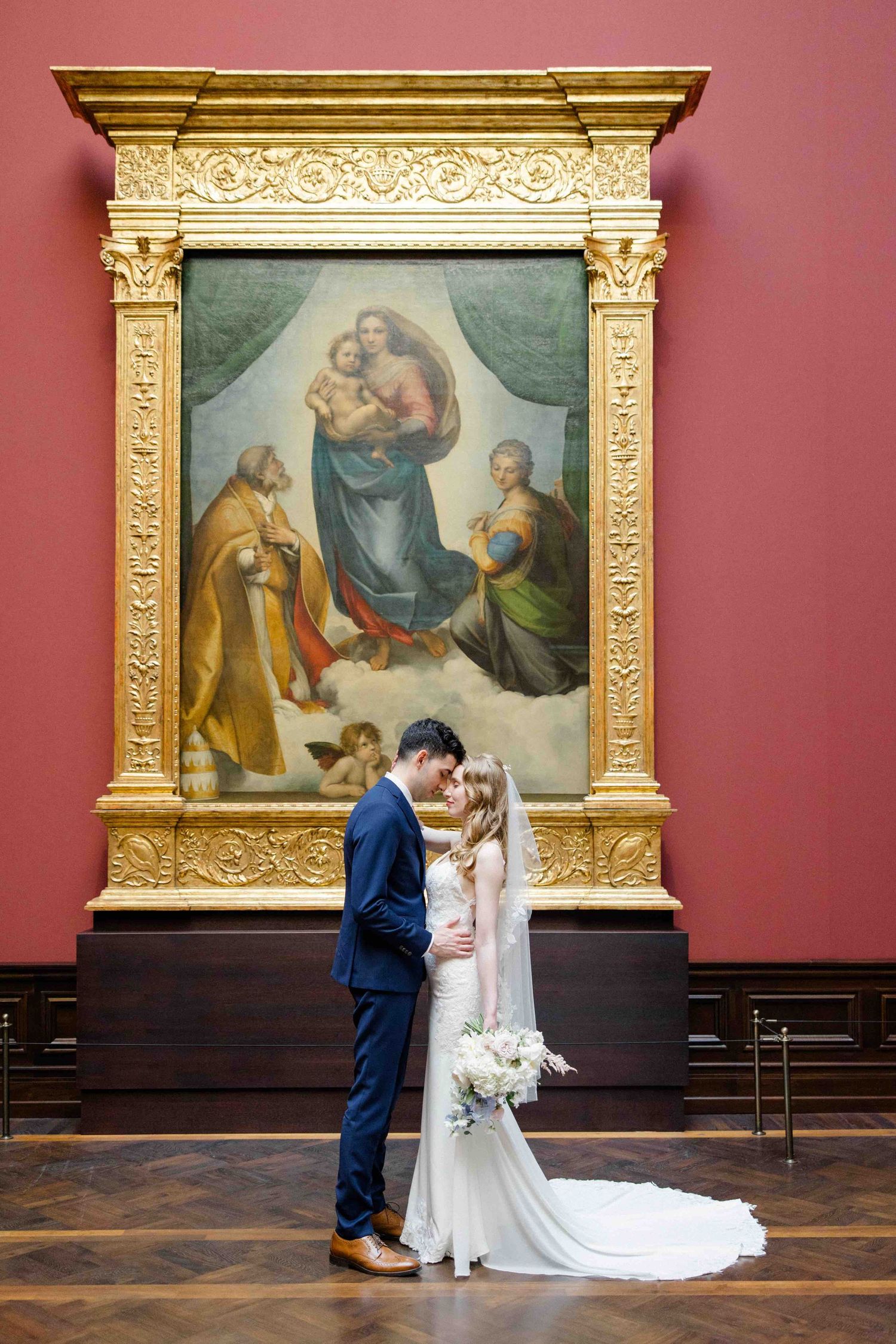 Wedding couple stands in ornate museum gallery with Renaissance paintings and gilded frames against burgundy walls.