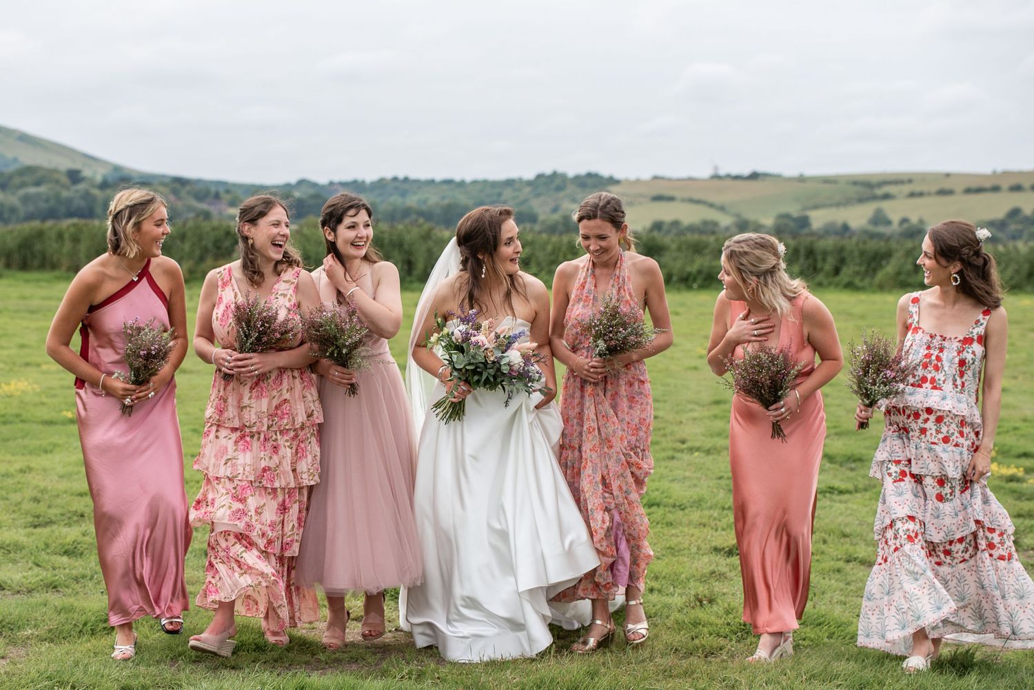 Bride and bridesmaids in various pink dresses walk through field laughing and looking at each other
