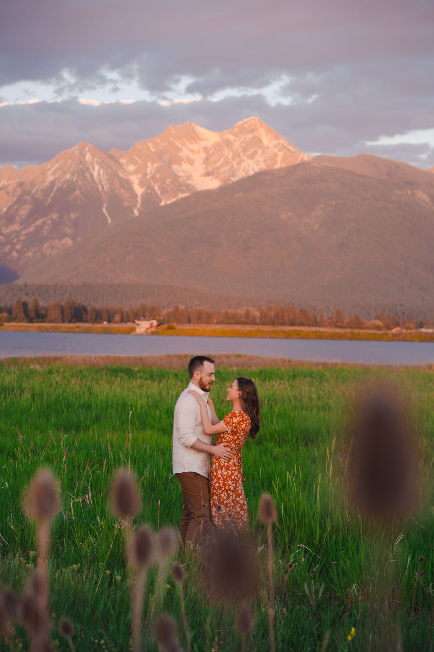 Mountain landscape at sunset with orange alpenglow illuminating snow-capped peaks behind a lush green meadow.