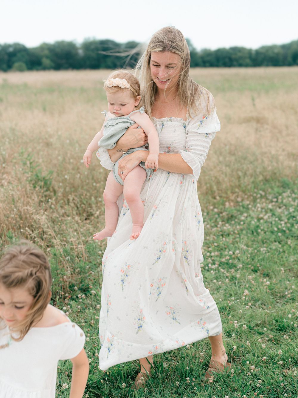 A mother wearing a doen dress holds her baby while walking in a grassy field in colts neck, New Jersey.