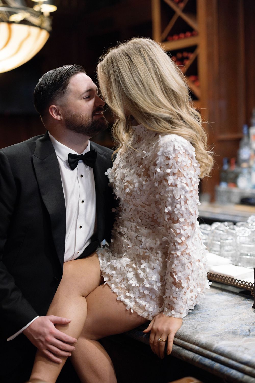 Romantic couple shares intimate moment at bar, bride wearing sparkly long sleeve mini dress and groom in black tuxedo.