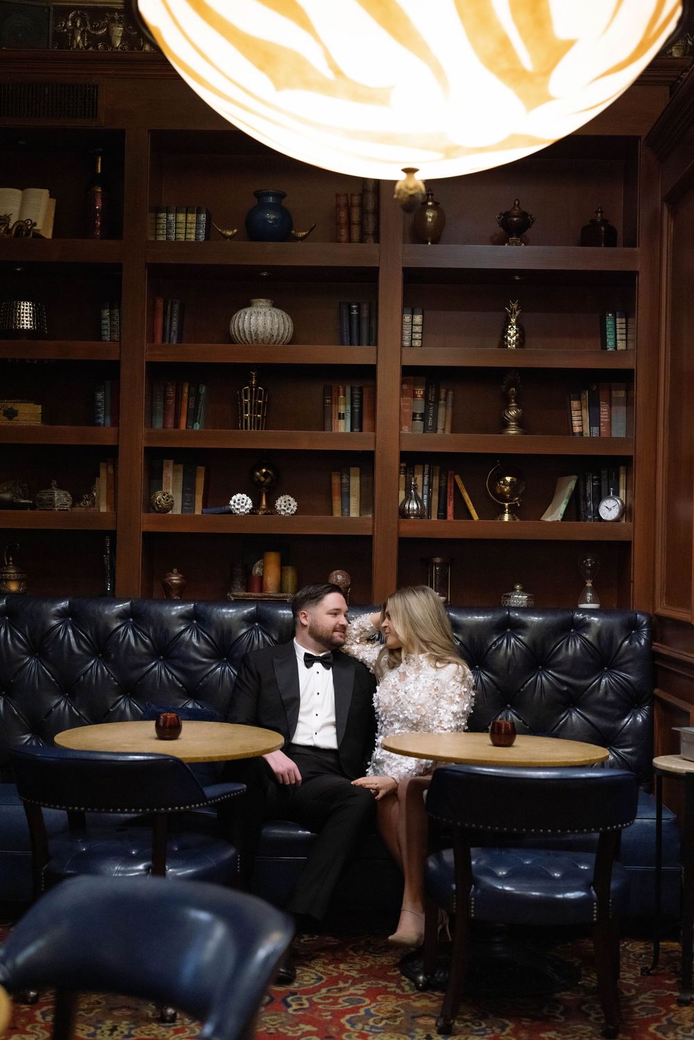 Elegant couple lounges on tufted leather banquette beneath vintage light fixture in grand library setting.